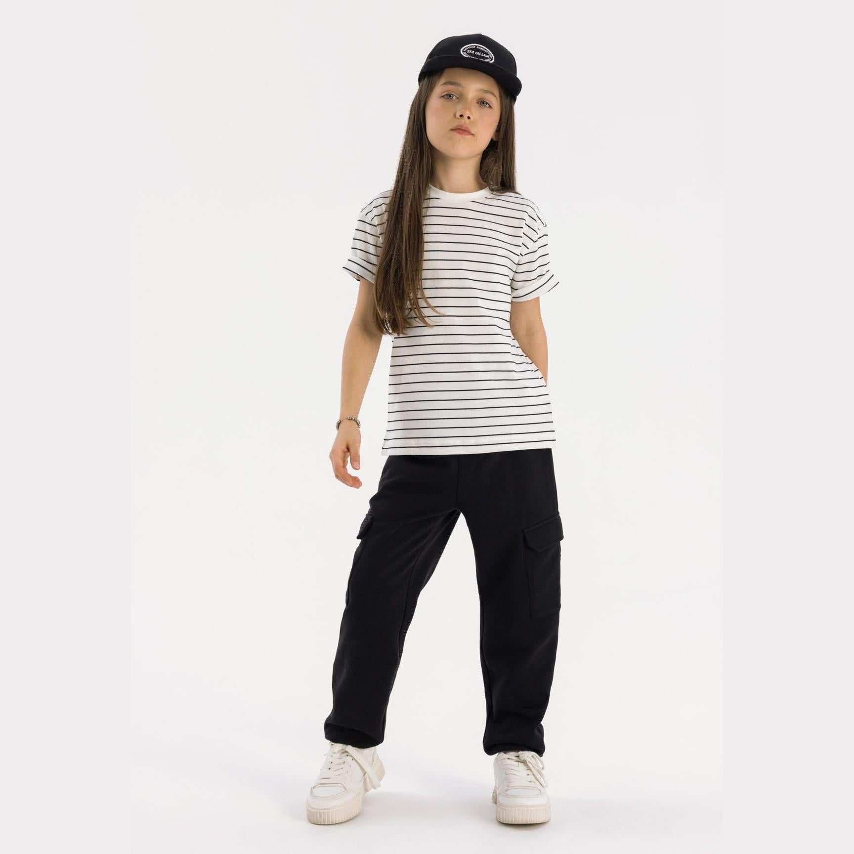 Girl wearing black fleece cargo joggers with white striped t-shirt and black cap, standing against white background