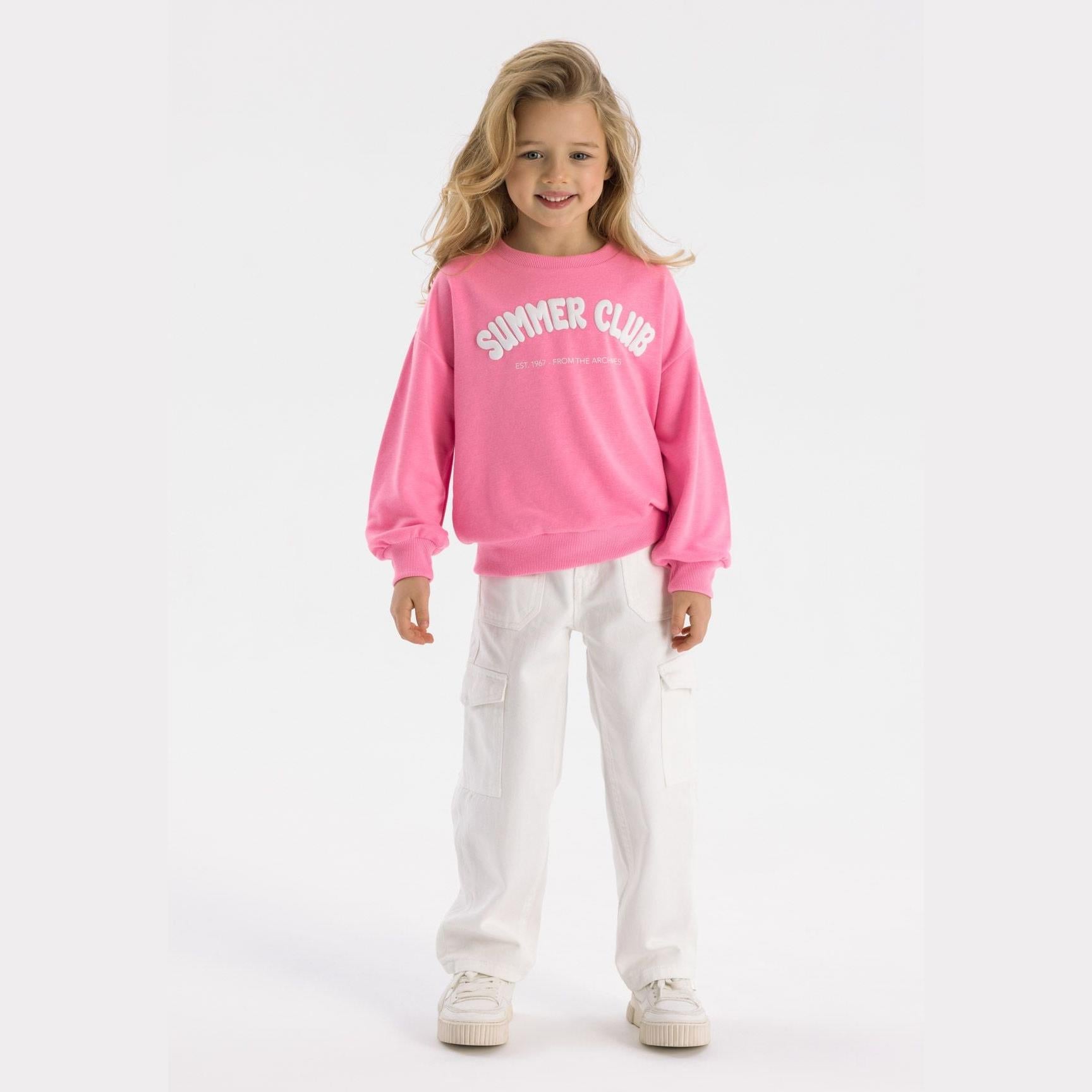Young girl wearing pink fleece sweatshirt with Summer Club print and white trousers, standing against white background