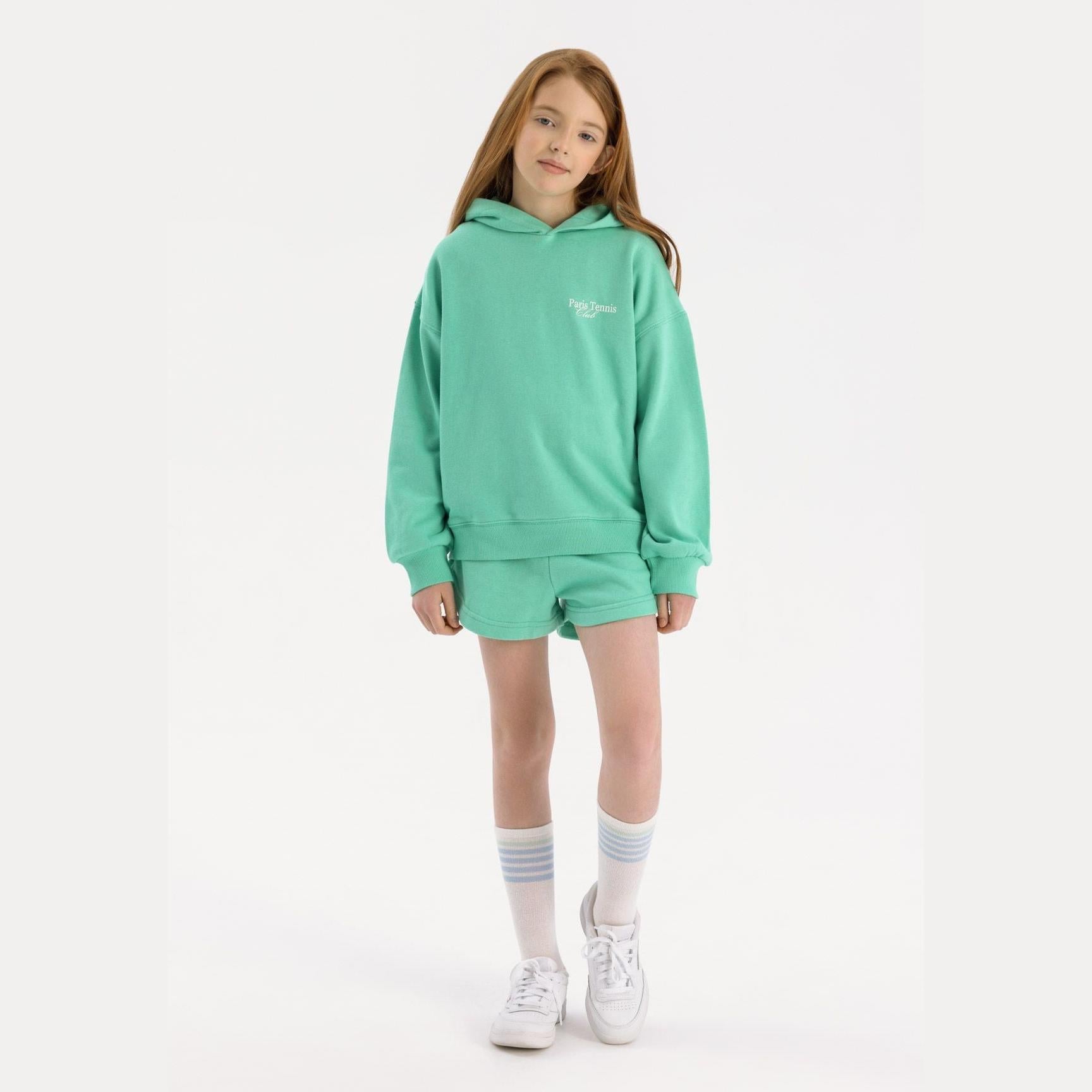 Young girl wearing green hoodie and shorts set against white background, styled with white sneakers and striped socks