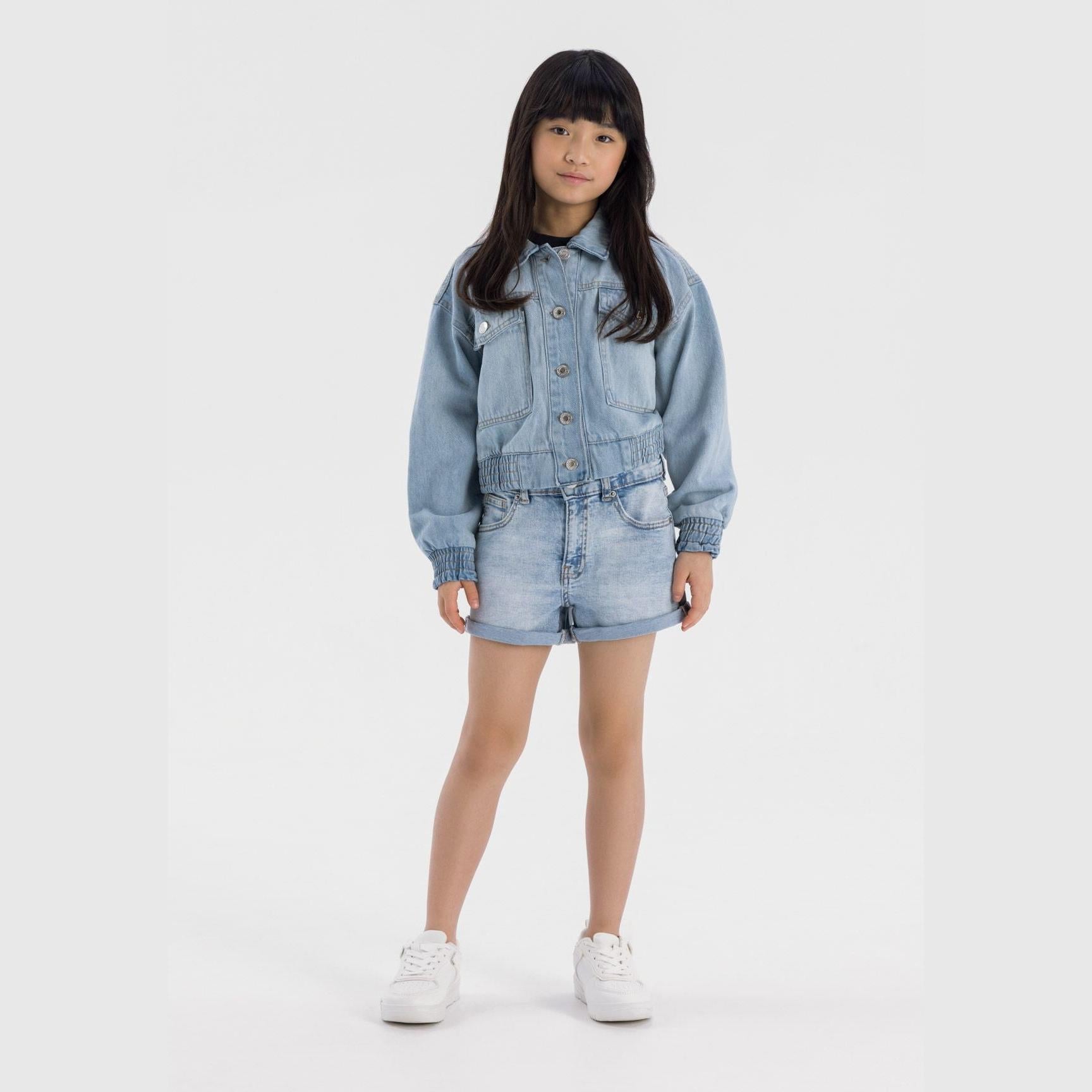 Girl wearing light blue cropped denim bomber jacket with matching shorts, white sneakers against white background, full body front view
