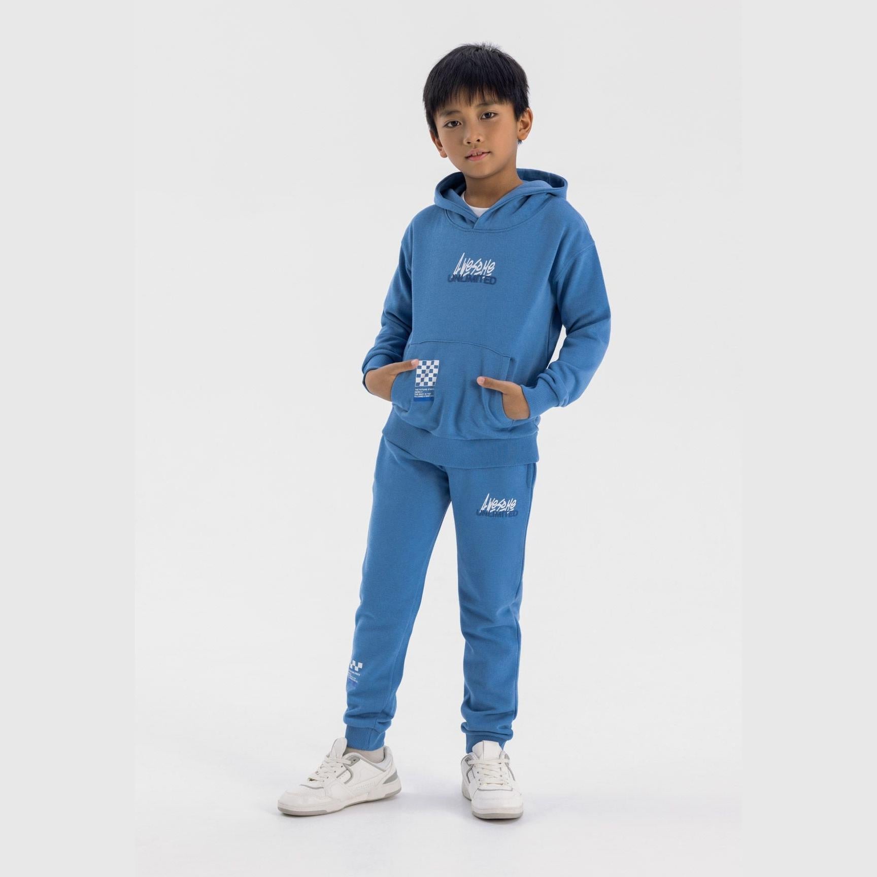 Young boy wearing blue fleece tracksuit set with Awesome Unlimited graphics, standing against white background with hands in hoodie pocket