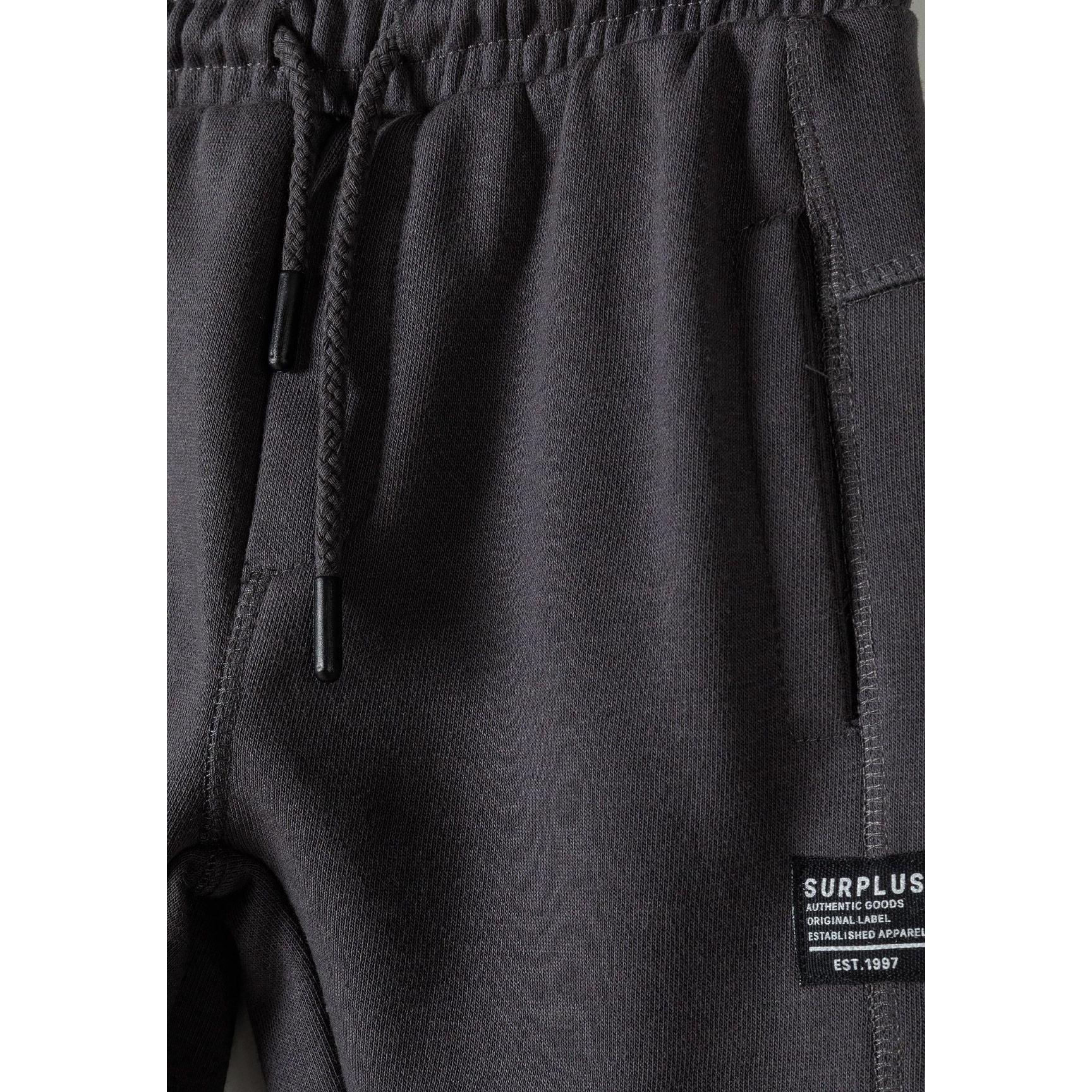 Close-up detail of grey joggers showing SURPLUS brand label, drawstring waist and panelled construction