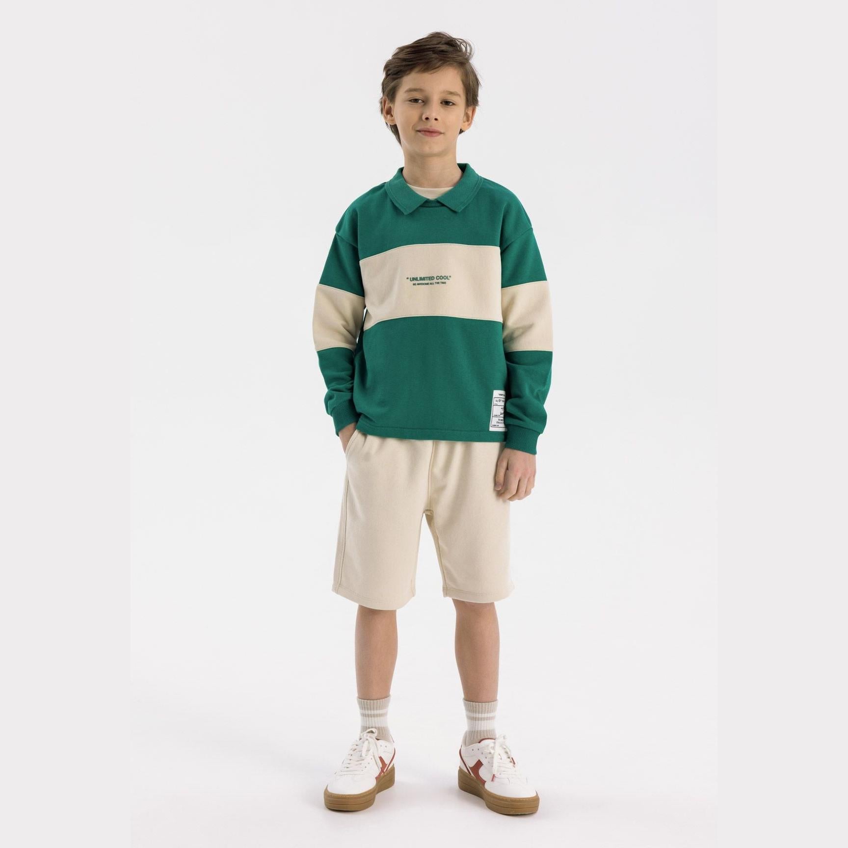Boy wearing green fleece polo jumper with cream contrast panel across chest, paired with cream shorts, standing against white background