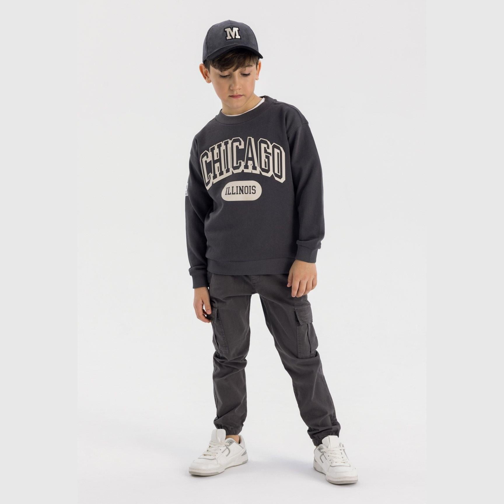 Boy wearing grey crew neck sweatshirt with Chicago Illinois print and matching cap, paired with grey cargo trousers and white trainers on light background