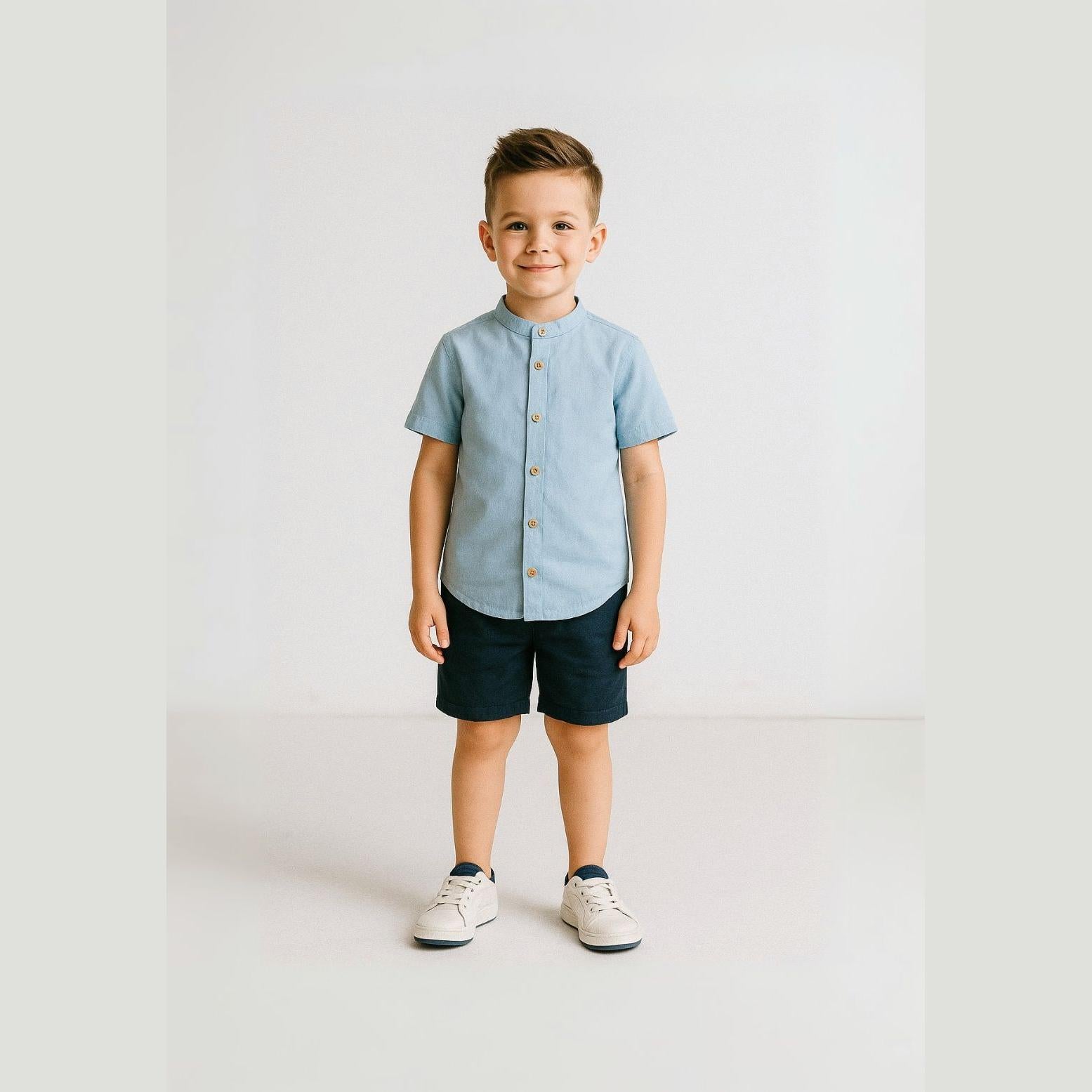 Young boy wearing light blue grandad collar shirt and navy shorts set, standing against white background, smiling at camera