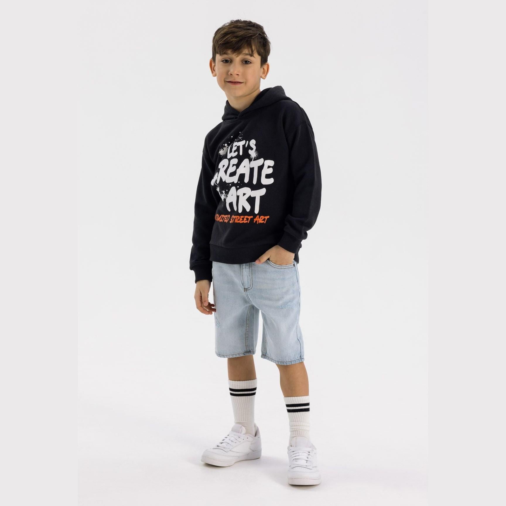 Boy wearing light blue ripped denim shorts with black hoodie and white sneakers, standing pose on white background