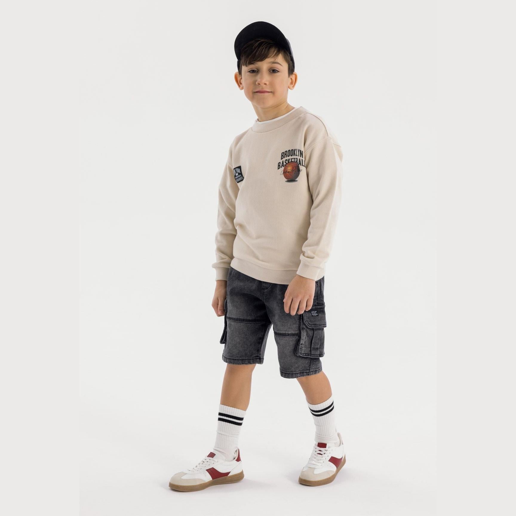 Young boy wearing beige basketball sweatshirt and black denim cargo shorts with white striped socks and red-white trainers against light grey background