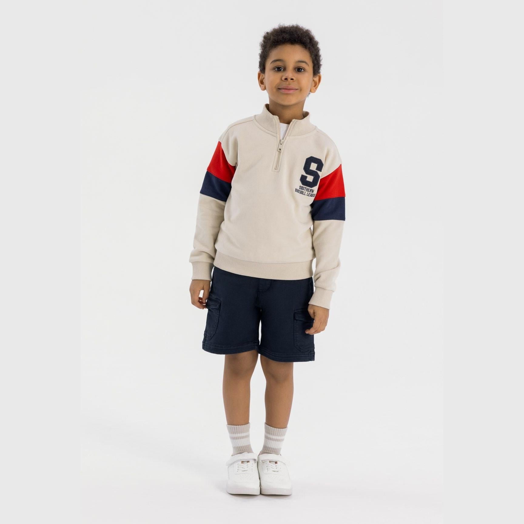 Young boy wearing navy cargo shorts with beige sweatshirt featuring red and navy sleeve details, standing against white background
