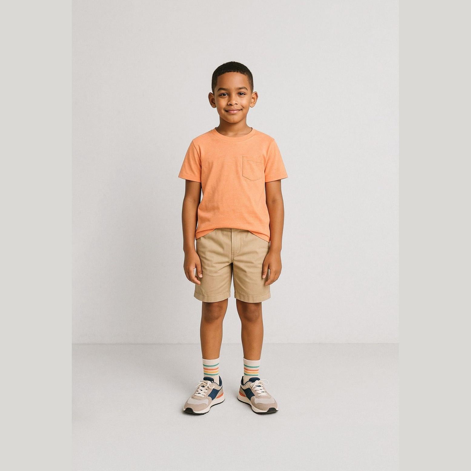Young boy wearing beige chino shorts with coral orange t-shirt, standing against neutral background in casual summer outfit