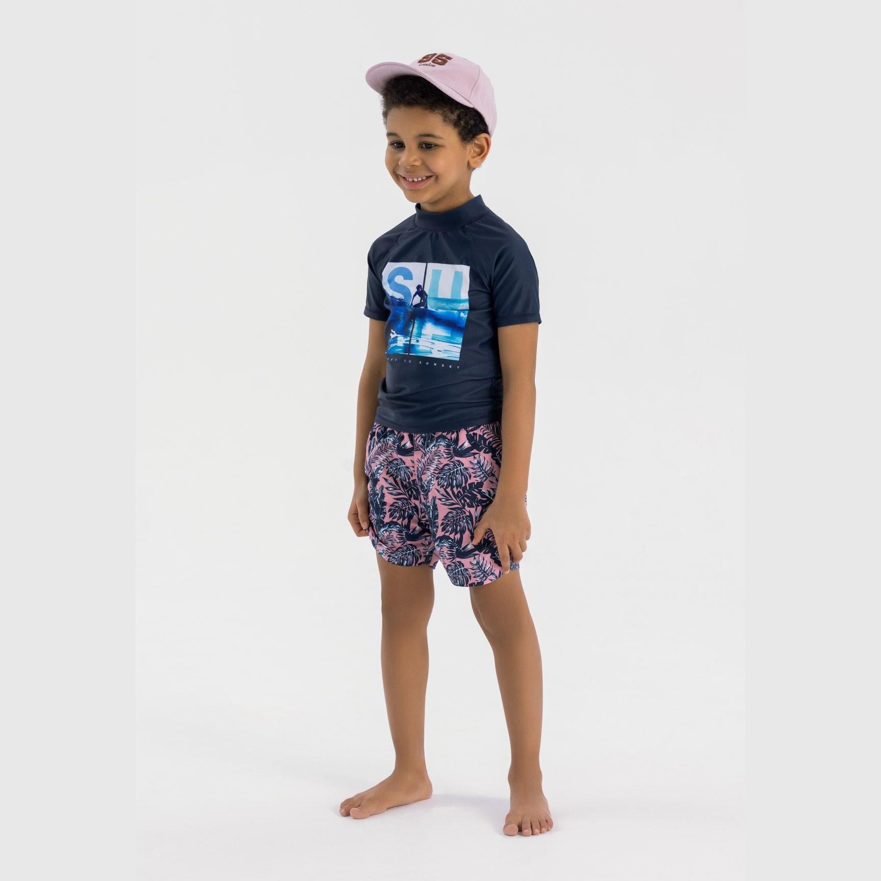 Young boy wearing navy swim top with surf print and patterned swim shorts, paired with pink cap, standing on white background