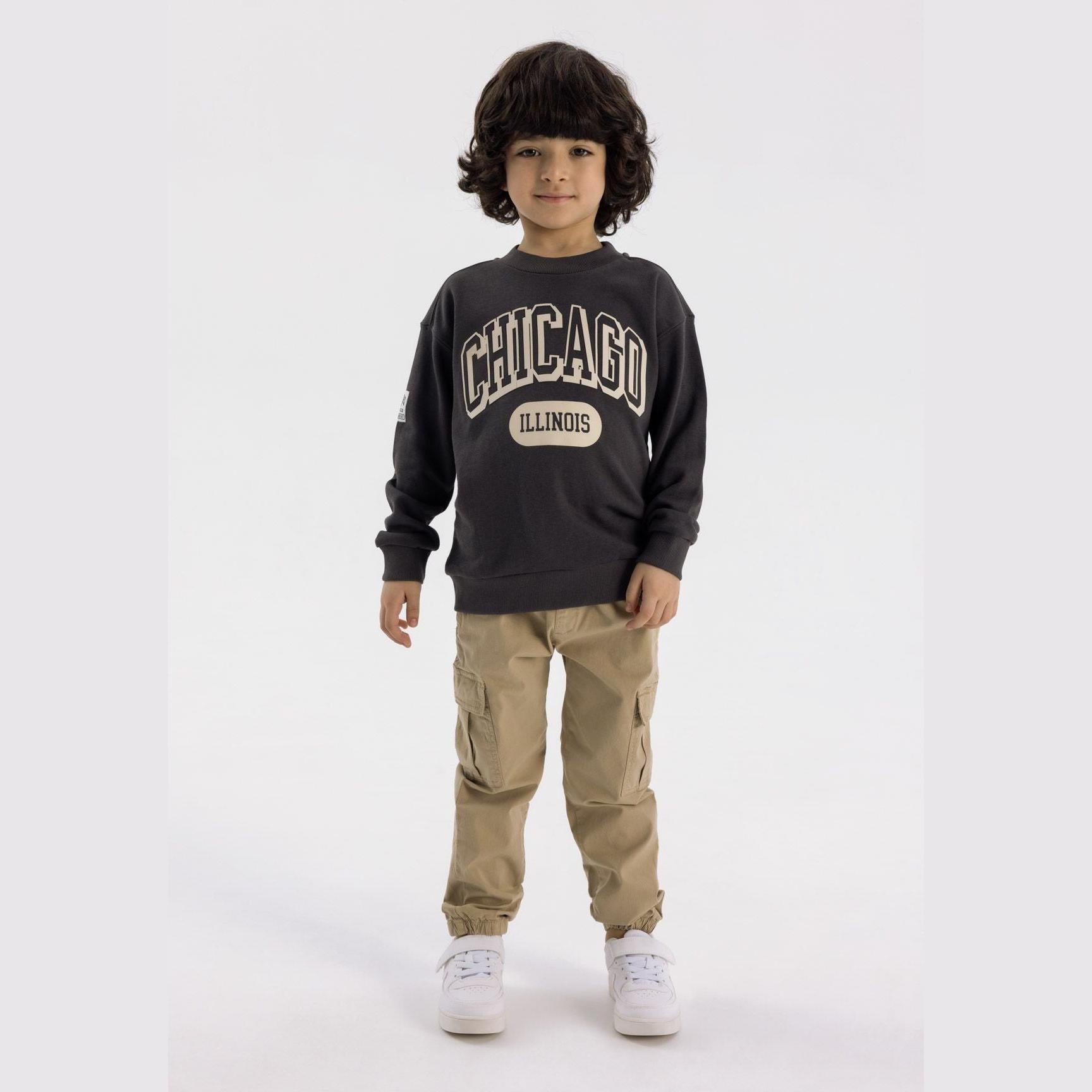 Young boy wearing khaki cargo trousers with dark sweatshirt, standing against white background, front view showing full outfit