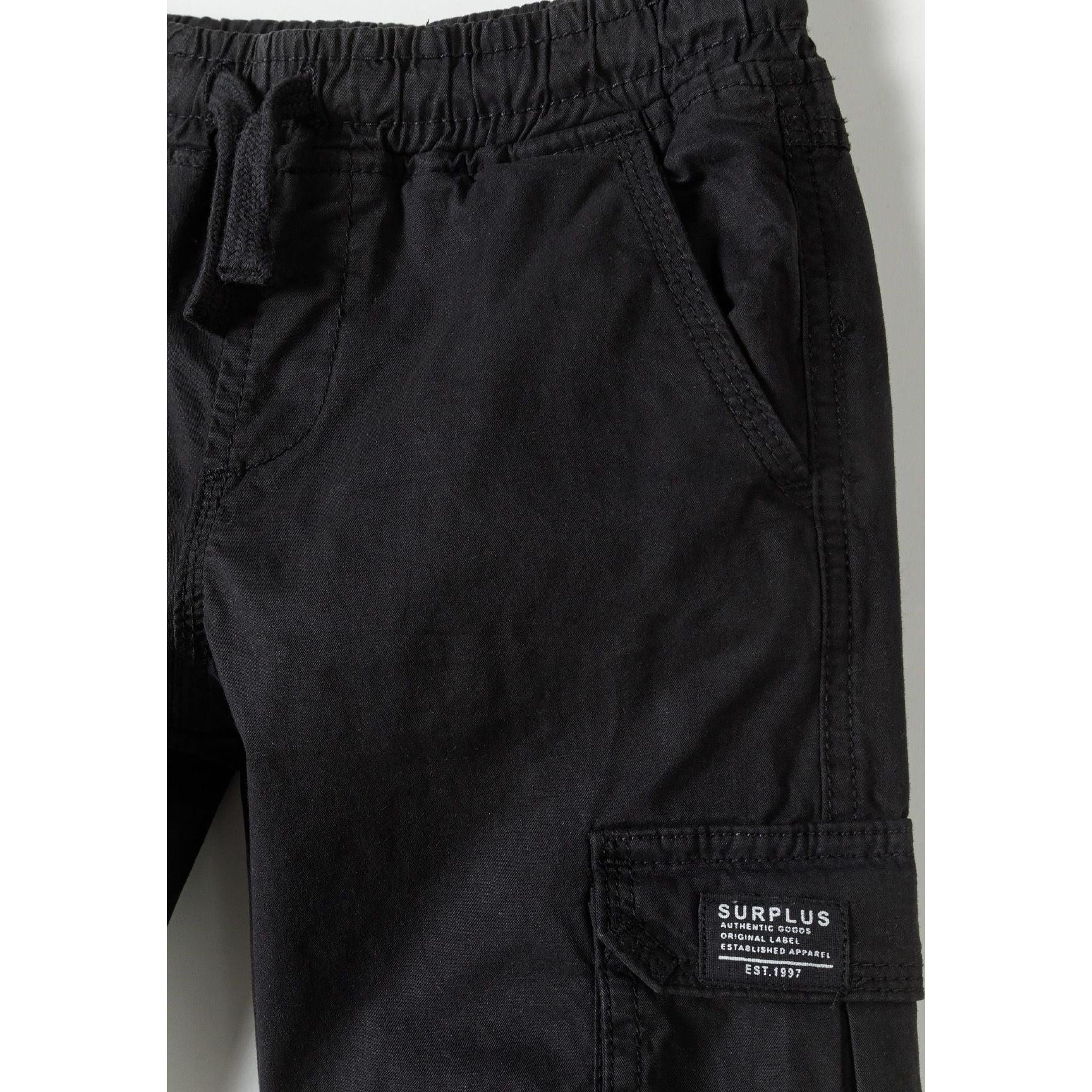 Close-up detail of black cargo trousers showing SURPLUS branded cargo pocket, elasticated waistband with drawcord and fabric texture