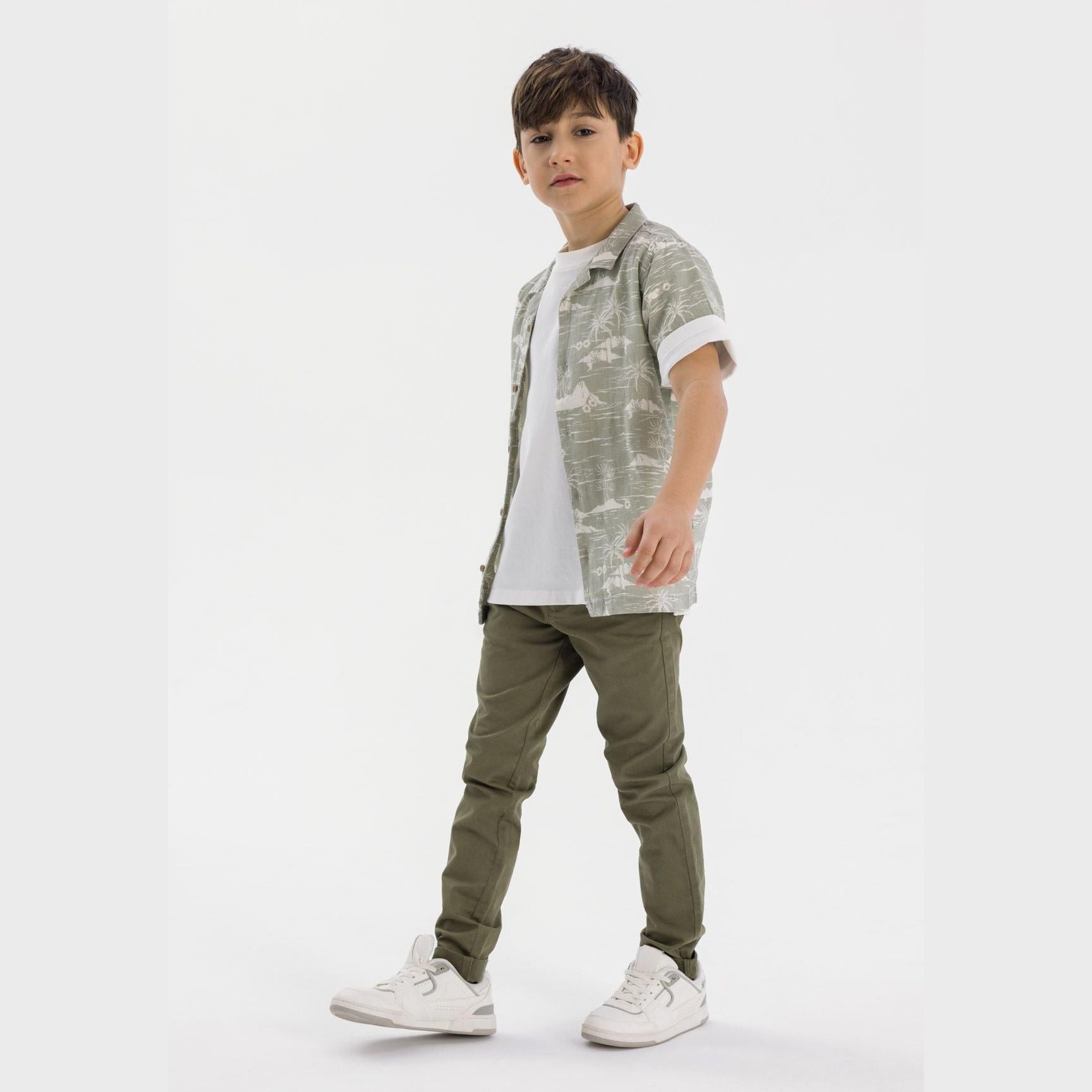 Young boy wearing olive chino trousers with short-sleeve shirt and white trainers, standing against white studio background