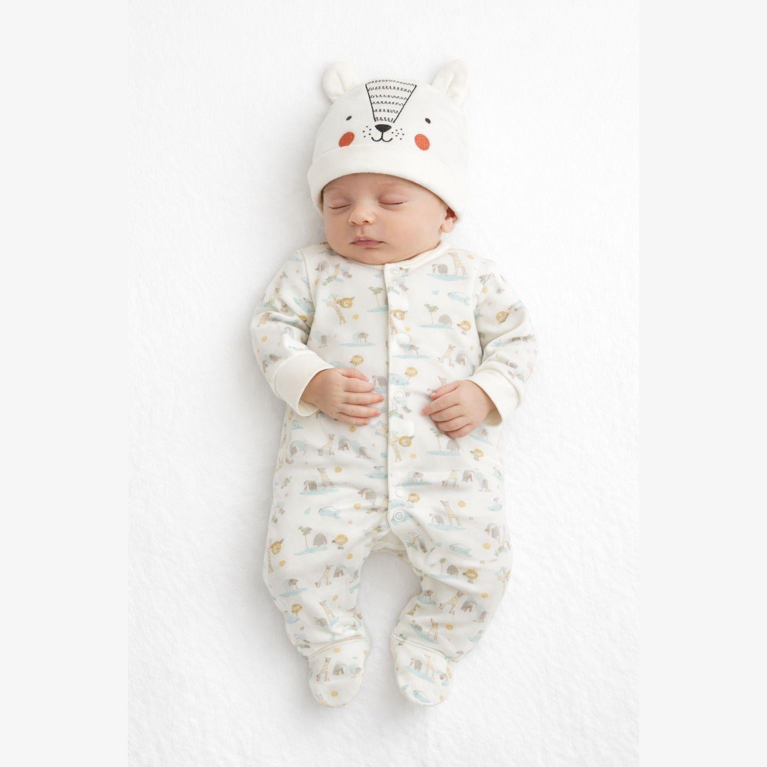 Baby wearing cream cotton sleepsuit with bear print pattern and matching bear face hat with ears, lying on white background