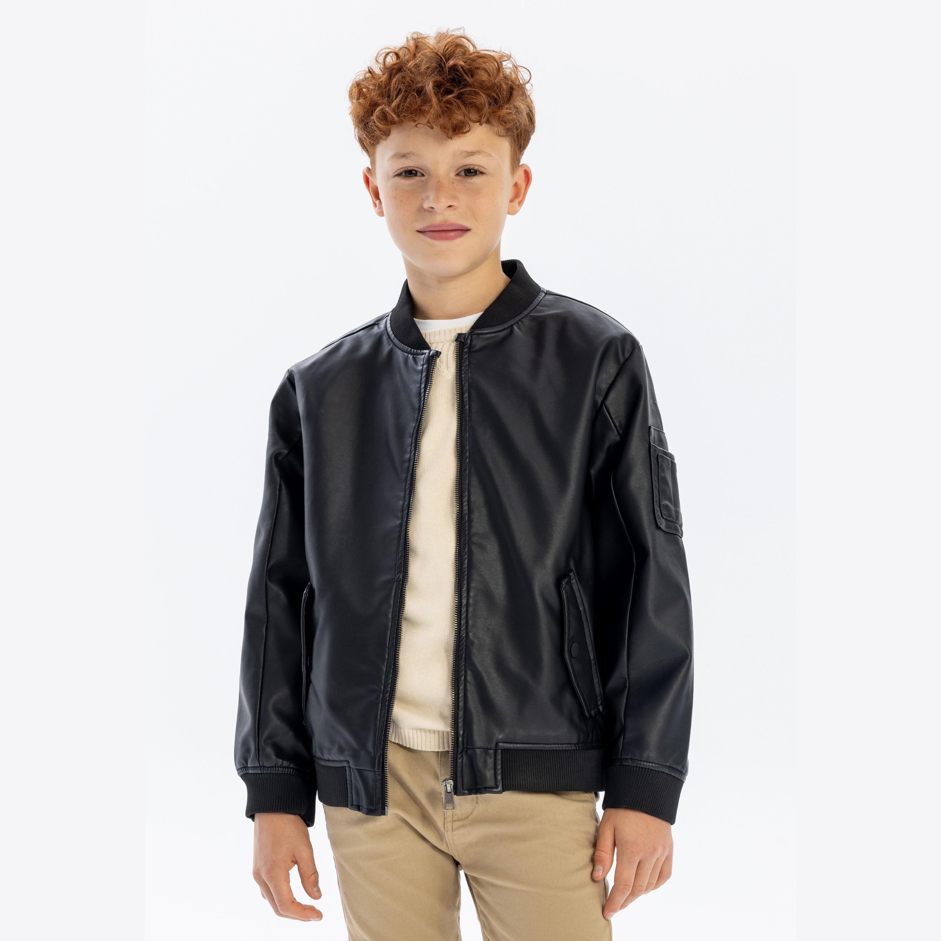 Young boy with curly hair wearing black faux leather bomber jacket over cream sweater and beige trousers, standing against white background