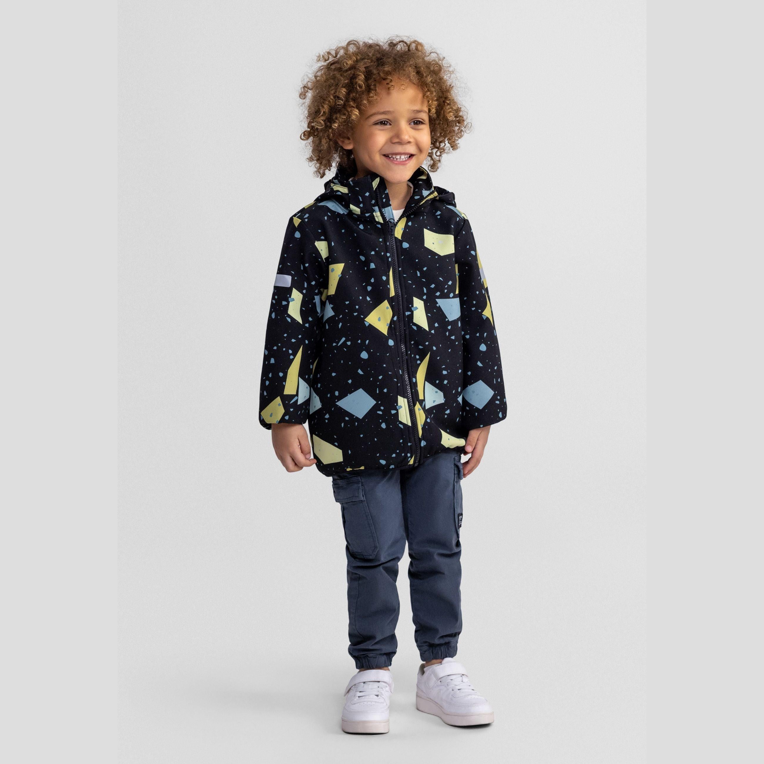 Young child with curly hair wearing gunmetal softshell jacket with abstract geometric print, paired with blue jeans and white sneakers, standing against light grey background
