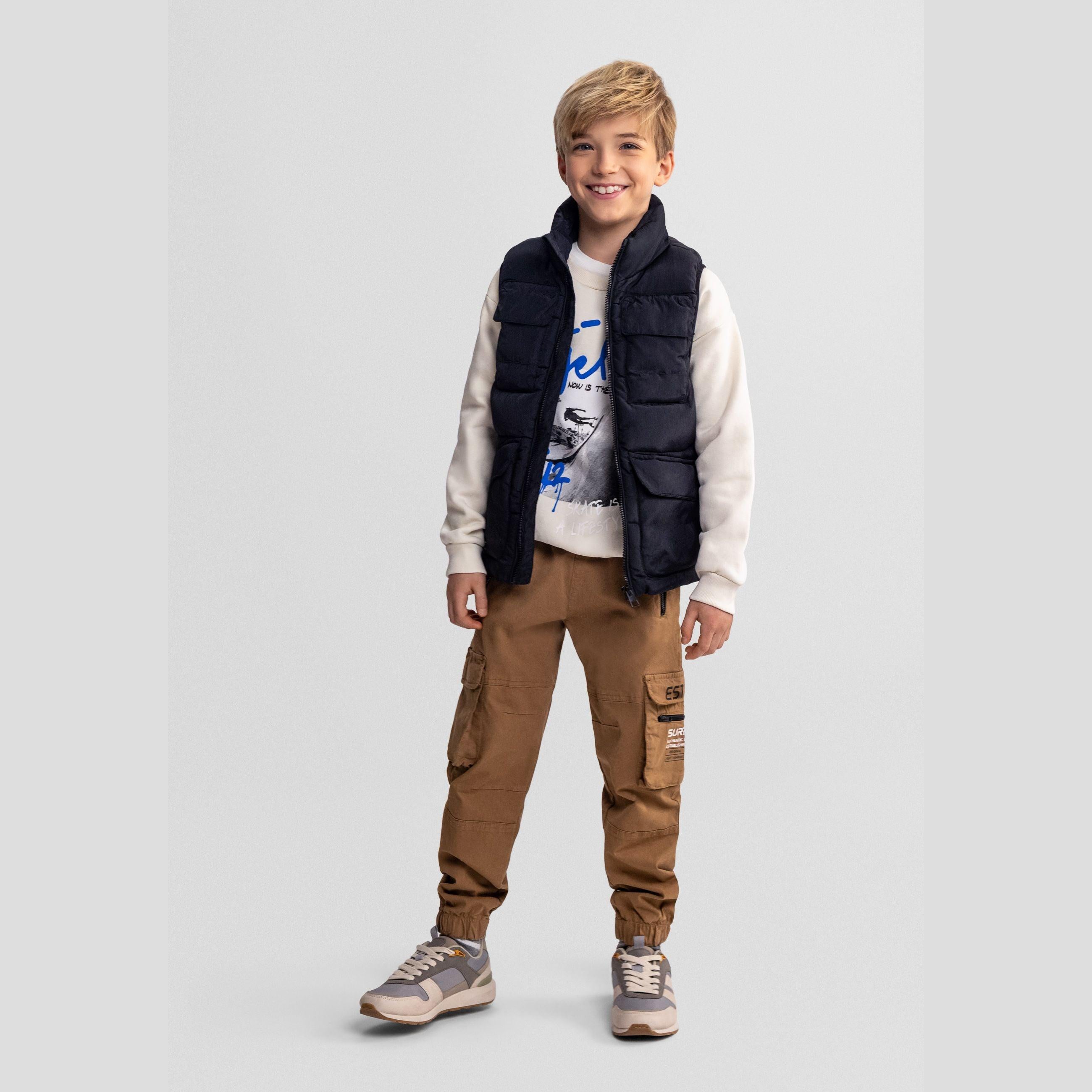 Young boy wearing black quilted waistcoat over white graphic sweatshirt and brown cargo pants, standing against light grey background