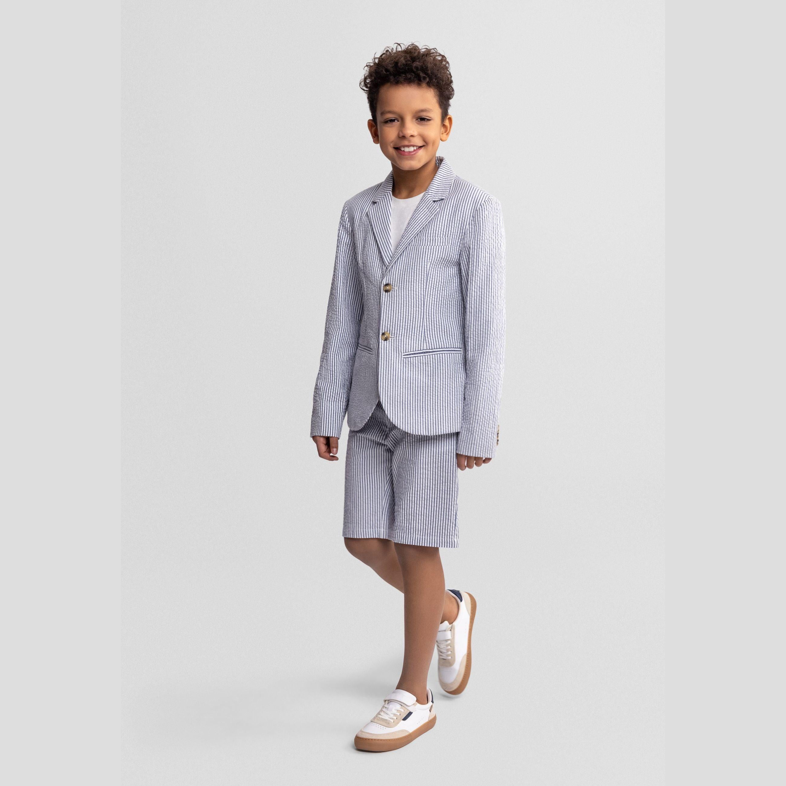 Boy wearing navy striped tailored blazer and matching shorts with white t-shirt and white sneakers, standing against light grey background