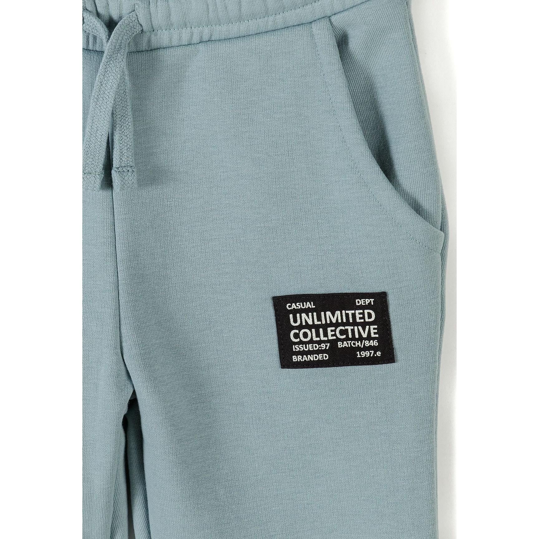 Close-up detail of sage blue jogger waistband and front pocket area showing black rectangular branded patch reading 'Unlimited Collective' with white text
