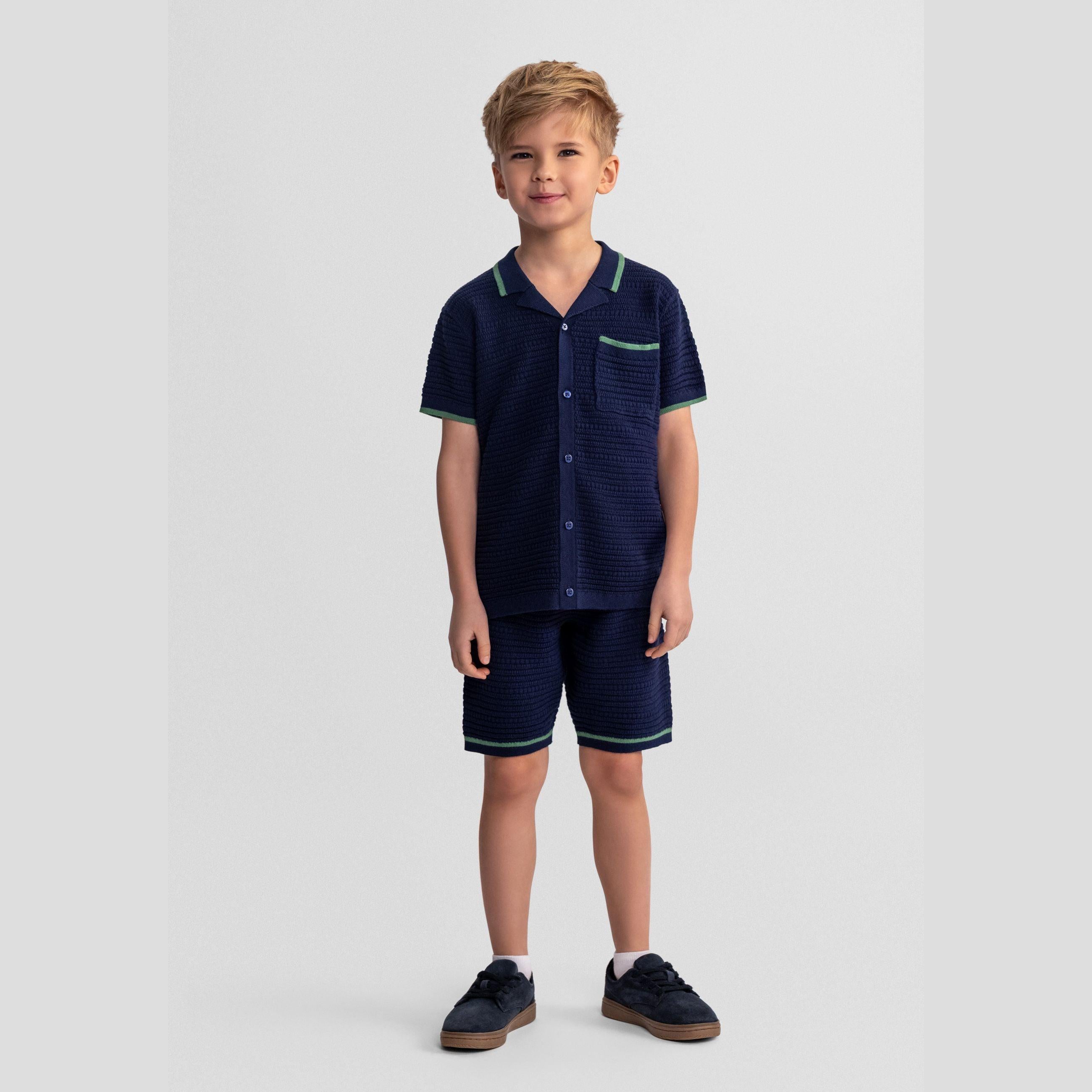 Young boy wearing navy blue knitted polo shirt and matching shorts set with green contrast trim, standing against white background