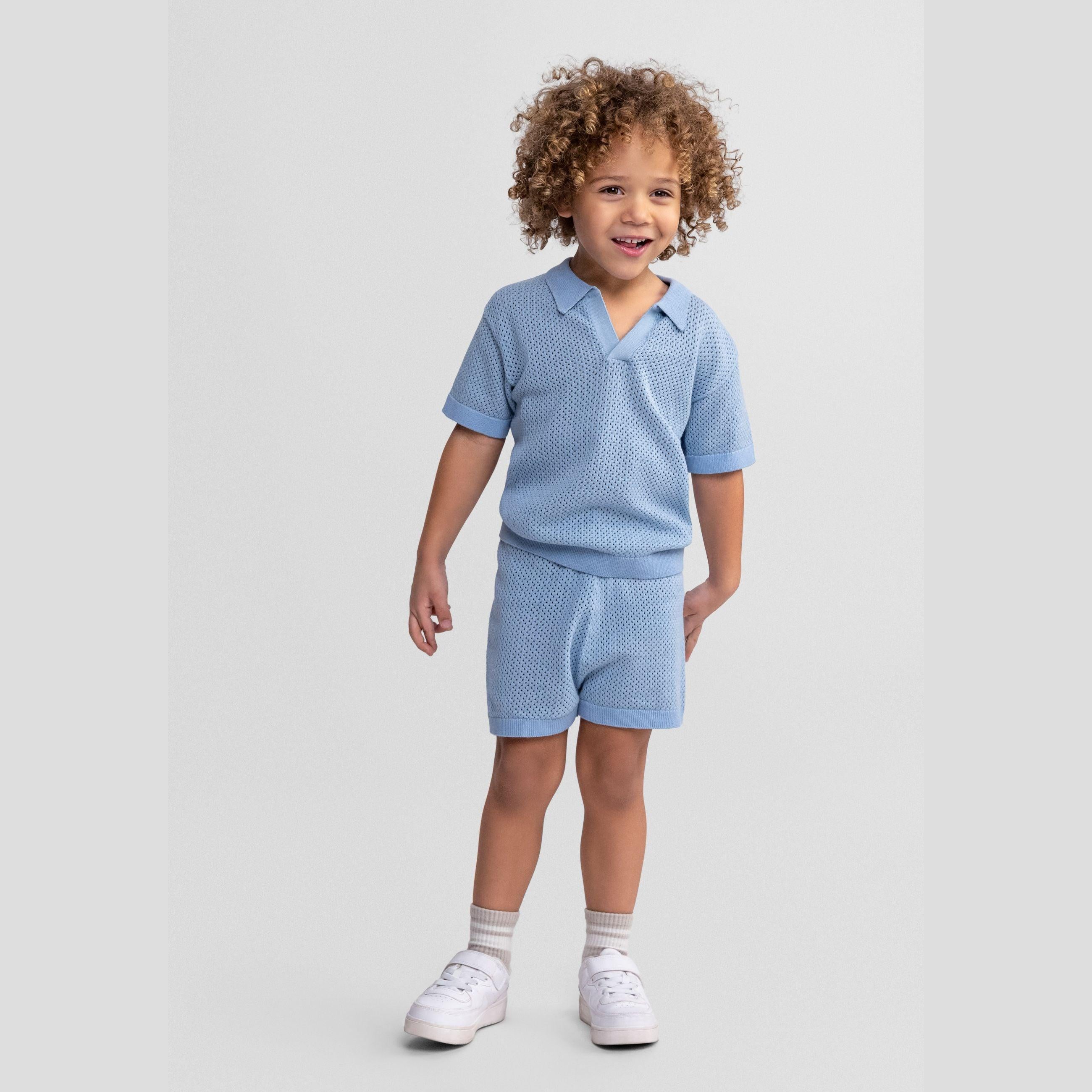 Child wearing light blue knitted polo shirt and matching shorts set, standing against grey background with curly hair and white sneakers