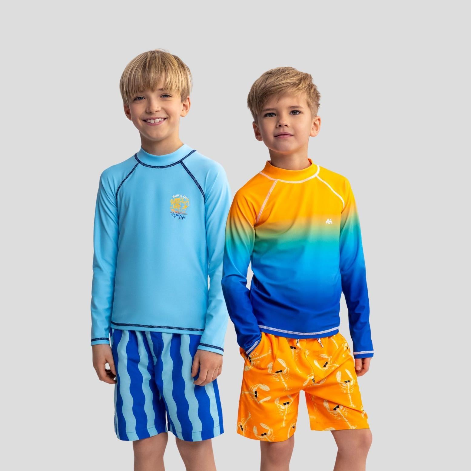 Two young boys wearing swim t-shirts and shorts standing against white background, one in solid blue with wave print shorts, one in orange-blue gradient with orange swim shorts