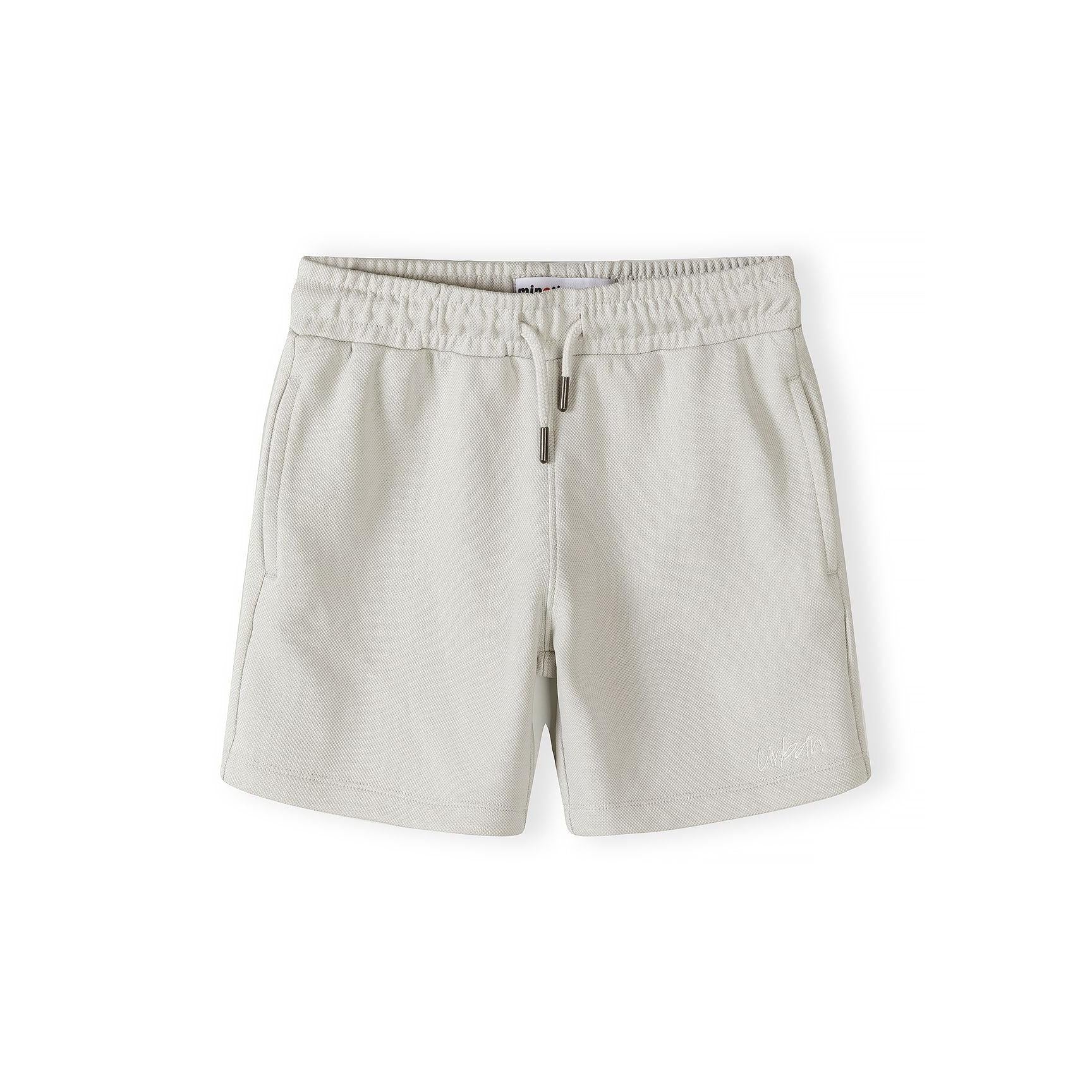 Close-up detail of grey pique polo shirt showing Minoti brand label, zip collar fastening and embroidered logo on textured fabric