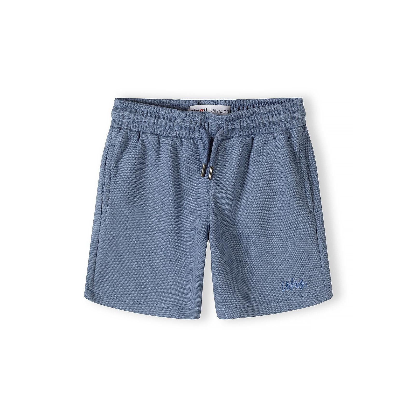 Close-up detail of blue polo shirt showing zip collar, embroidered Minoti logo, and pique texture with brand label visible