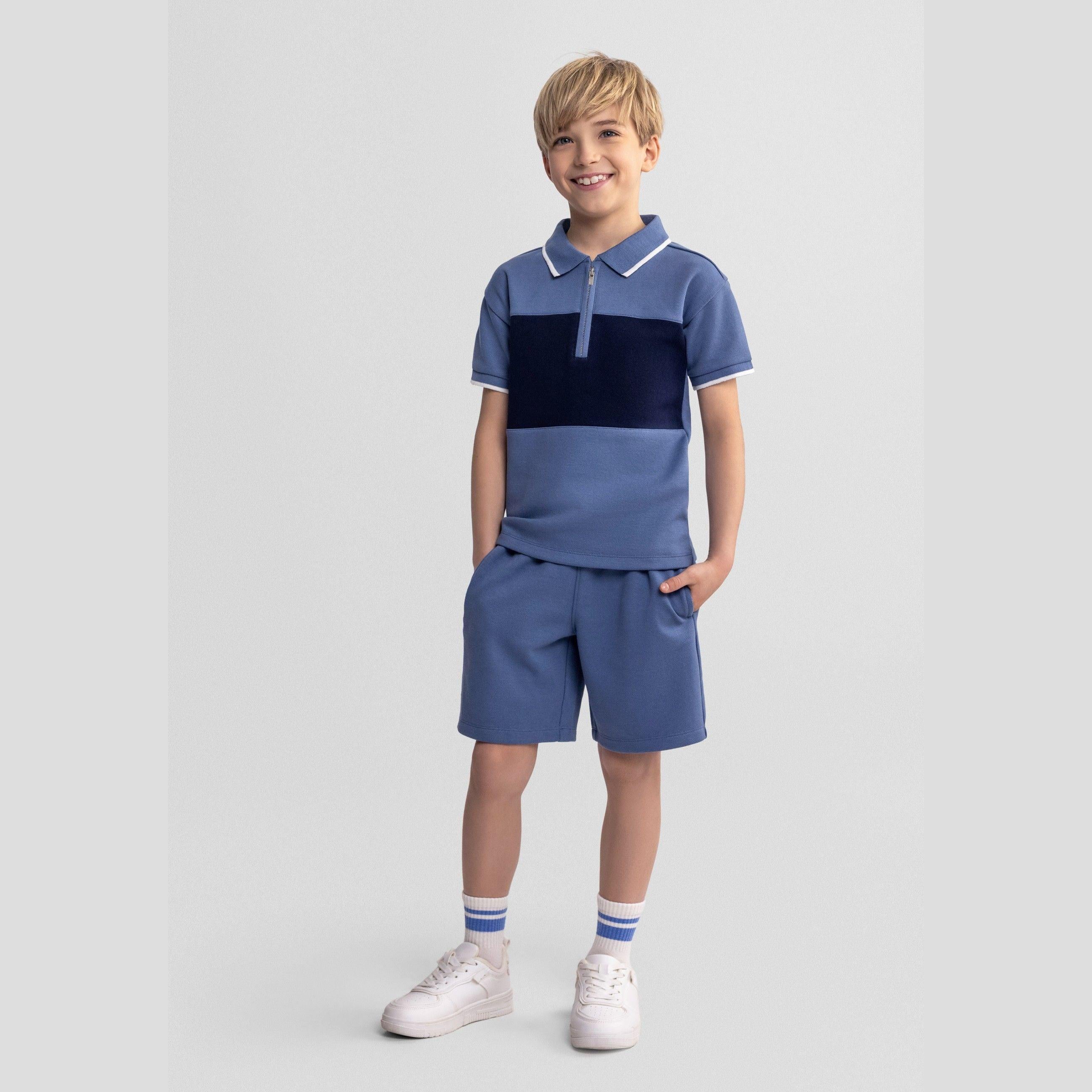 Young blonde boy wearing navy blue colour-blocked pique polo shirt with zip collar and matching shorts, standing against grey background with hands in pockets