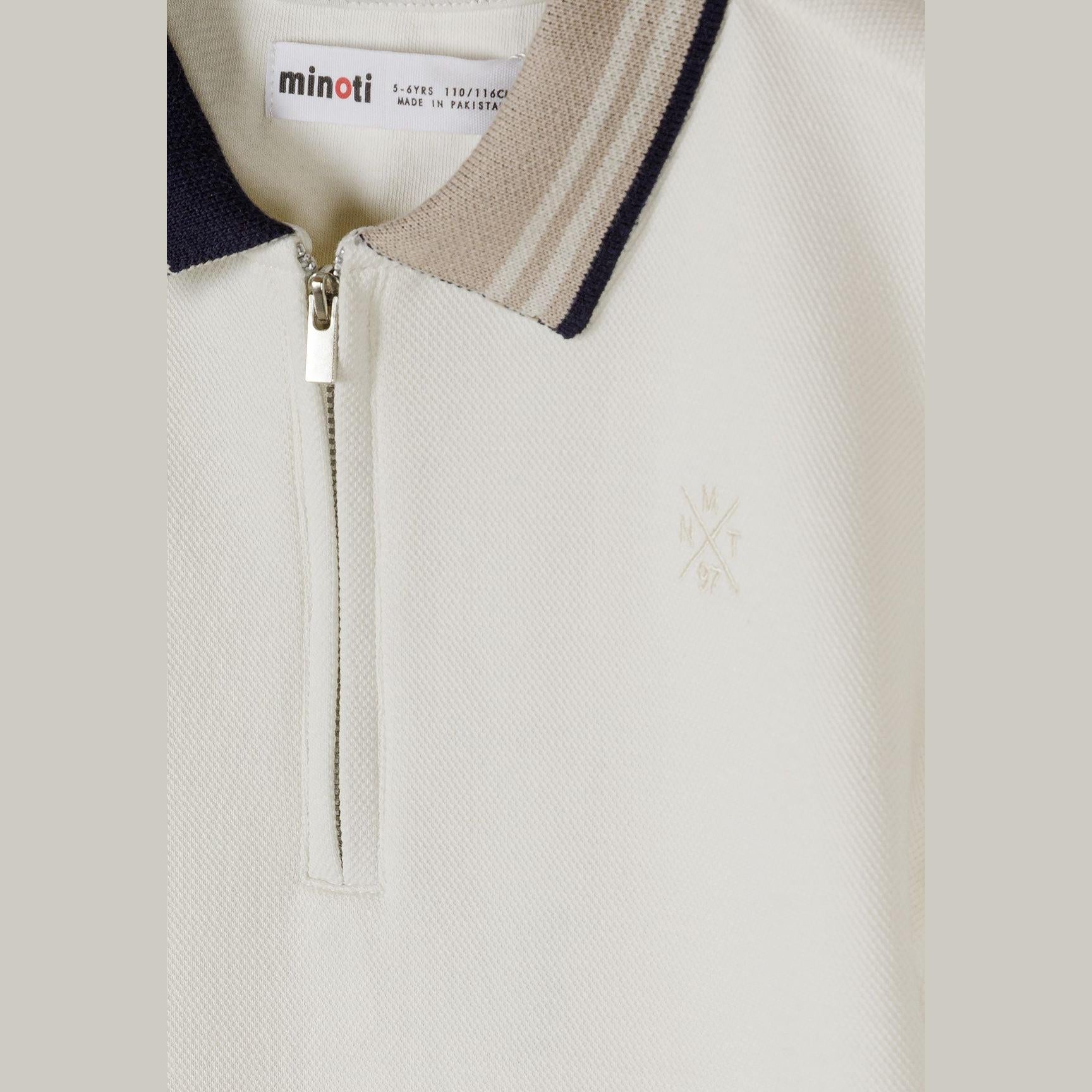 Close-up detail of cream pique polo shirt showing Minoti label, zip neck opening, navy collar and subtle embroidered logo on chest
