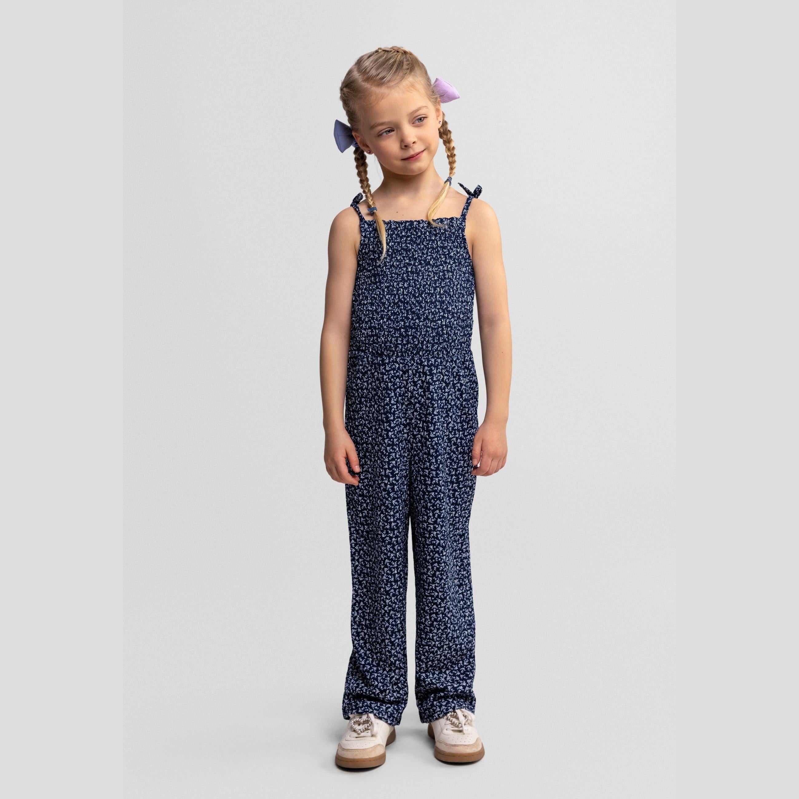 Young girl wearing navy floral jumpsuit with braided hair and hair bows, standing against light grey studio background, full body view showing complete outfit styling