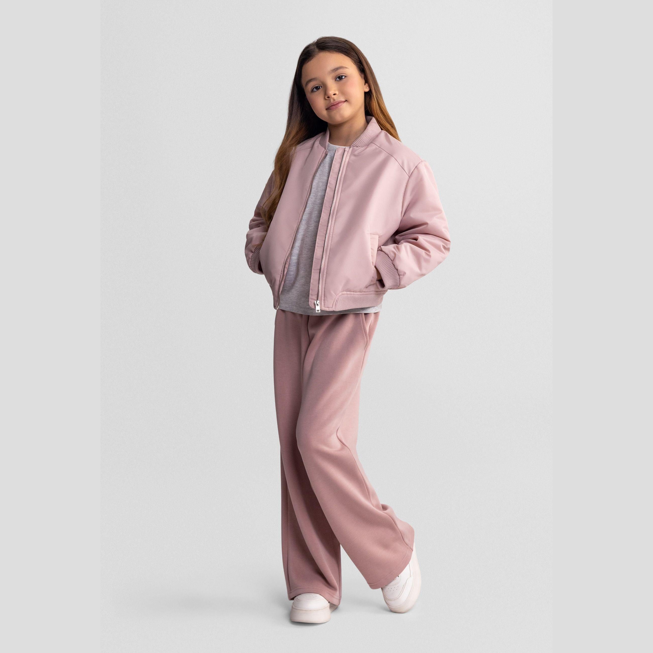 Girl wearing pink quilted bomber jacket with matching pink trousers, white sneakers, standing against white background, full body front view