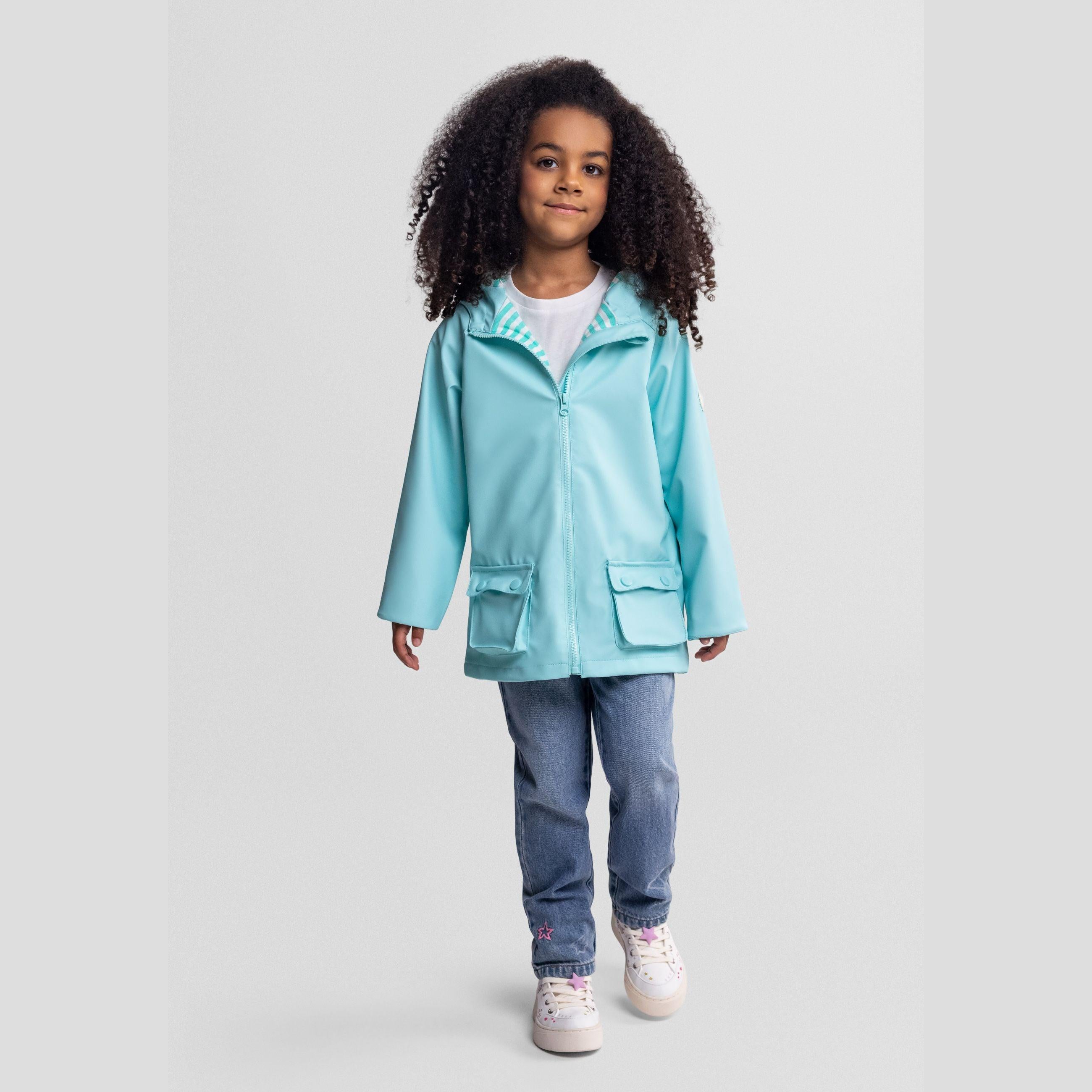 Young girl wearing mint green waterproof rain jacket with hood, paired with blue jeans and white sneakers, standing against light grey background