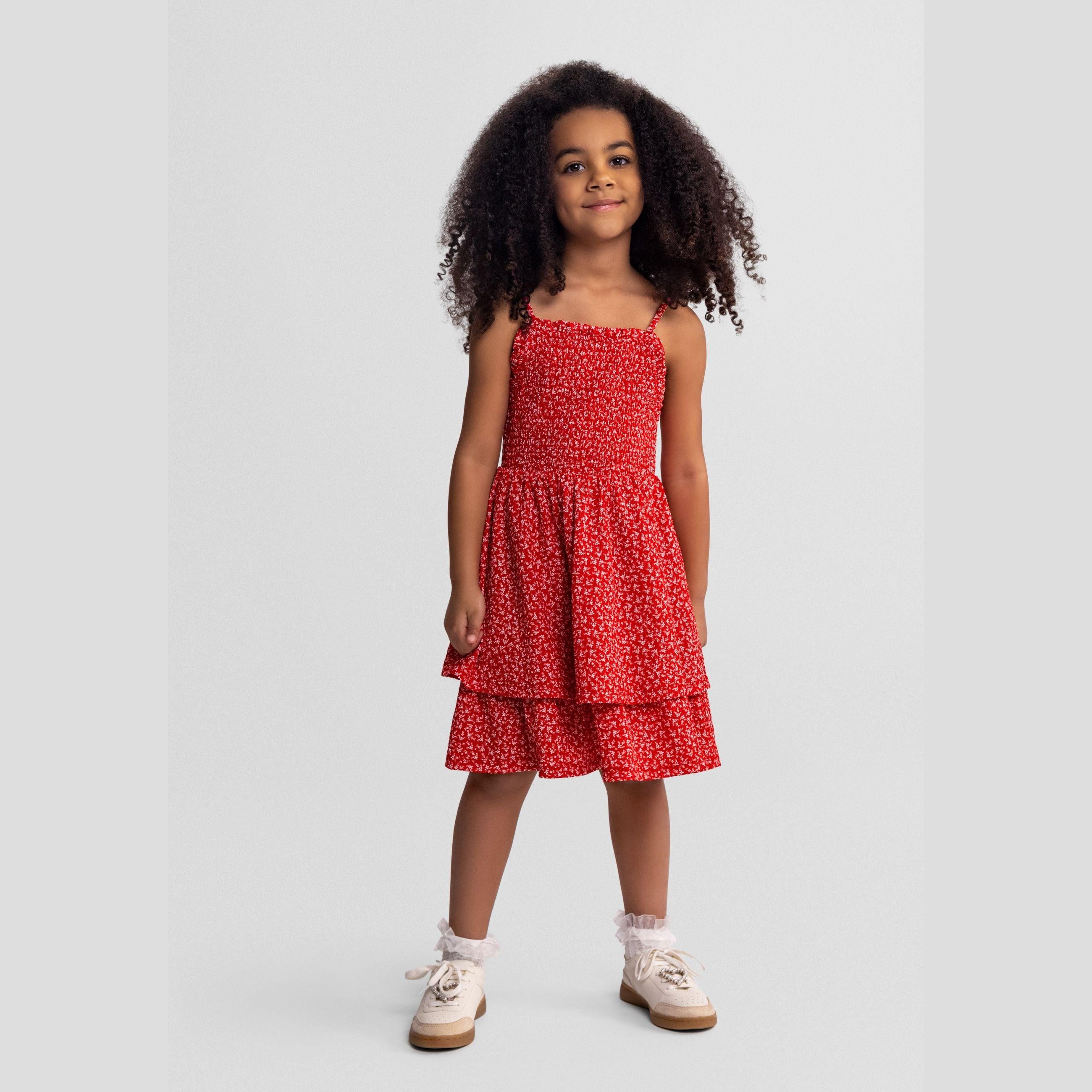 Young girl modeling red sleeveless dress with white floral print, featuring shirred bodice and tiered ruffle hem, standing against light grey background
