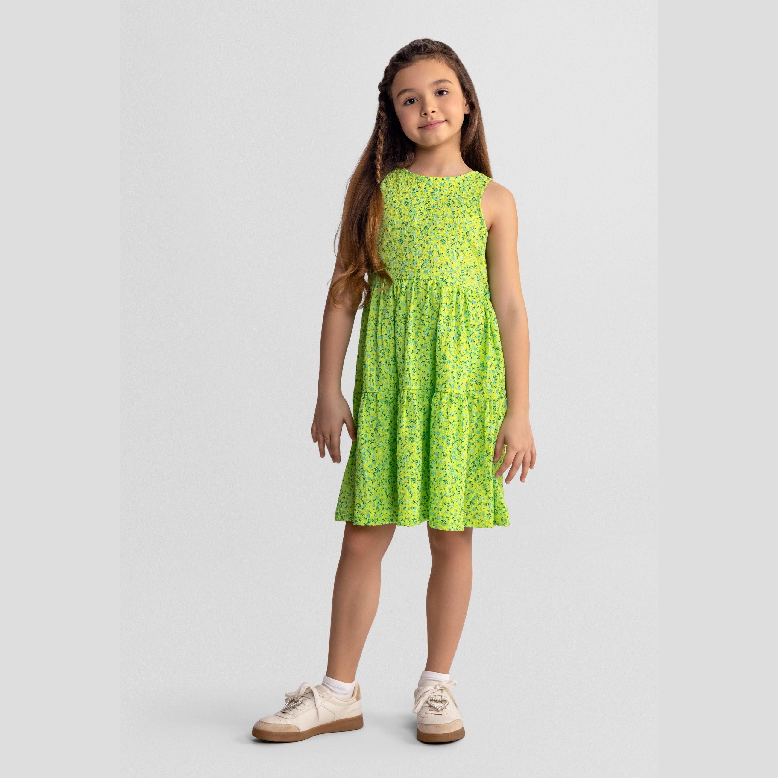 Young girl wearing emerald green floral sleeveless tiered dress, standing against light grey background, paired with cream canvas shoes