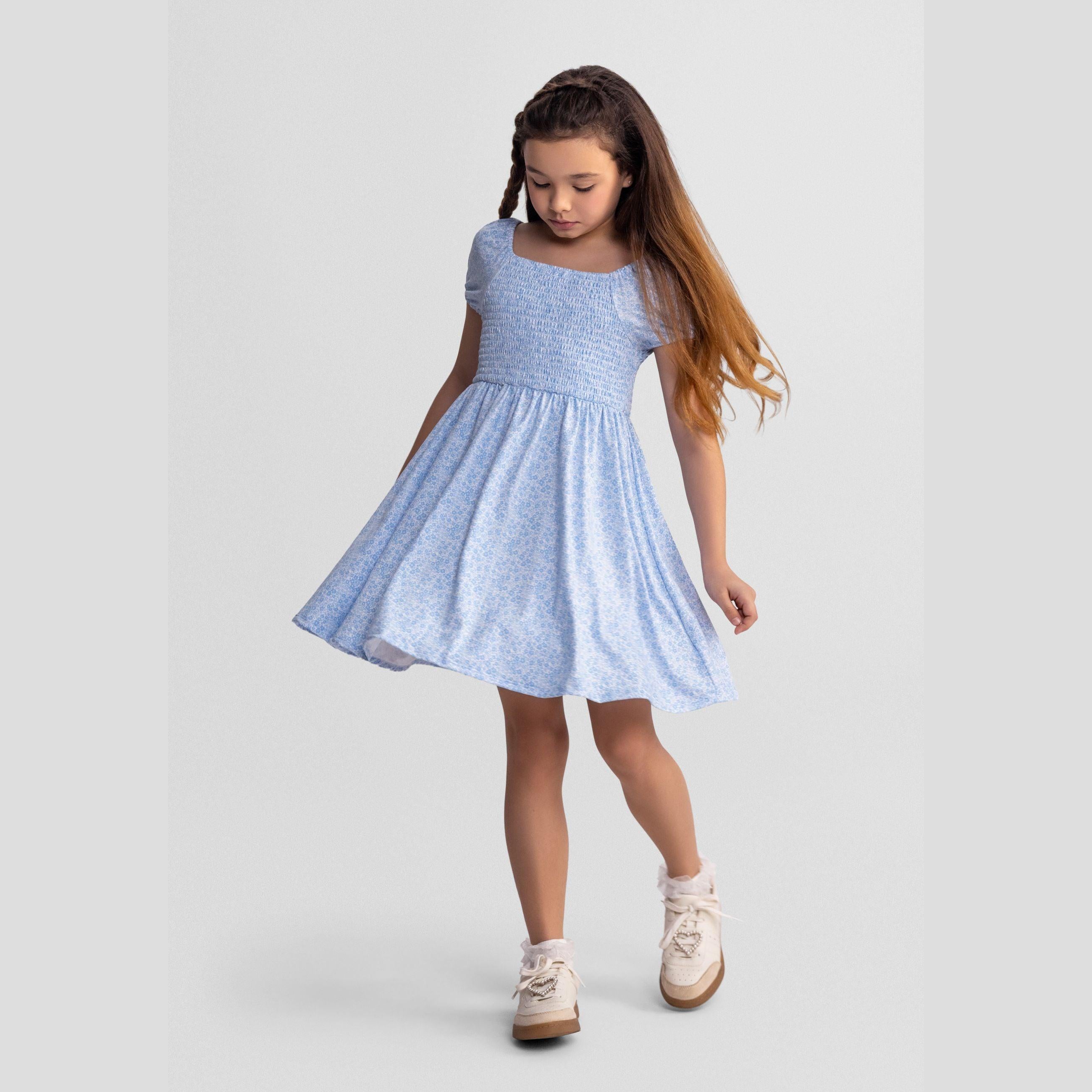 Young girl wearing light blue floral dress with square neckline and short sleeves, walking pose against white studio background