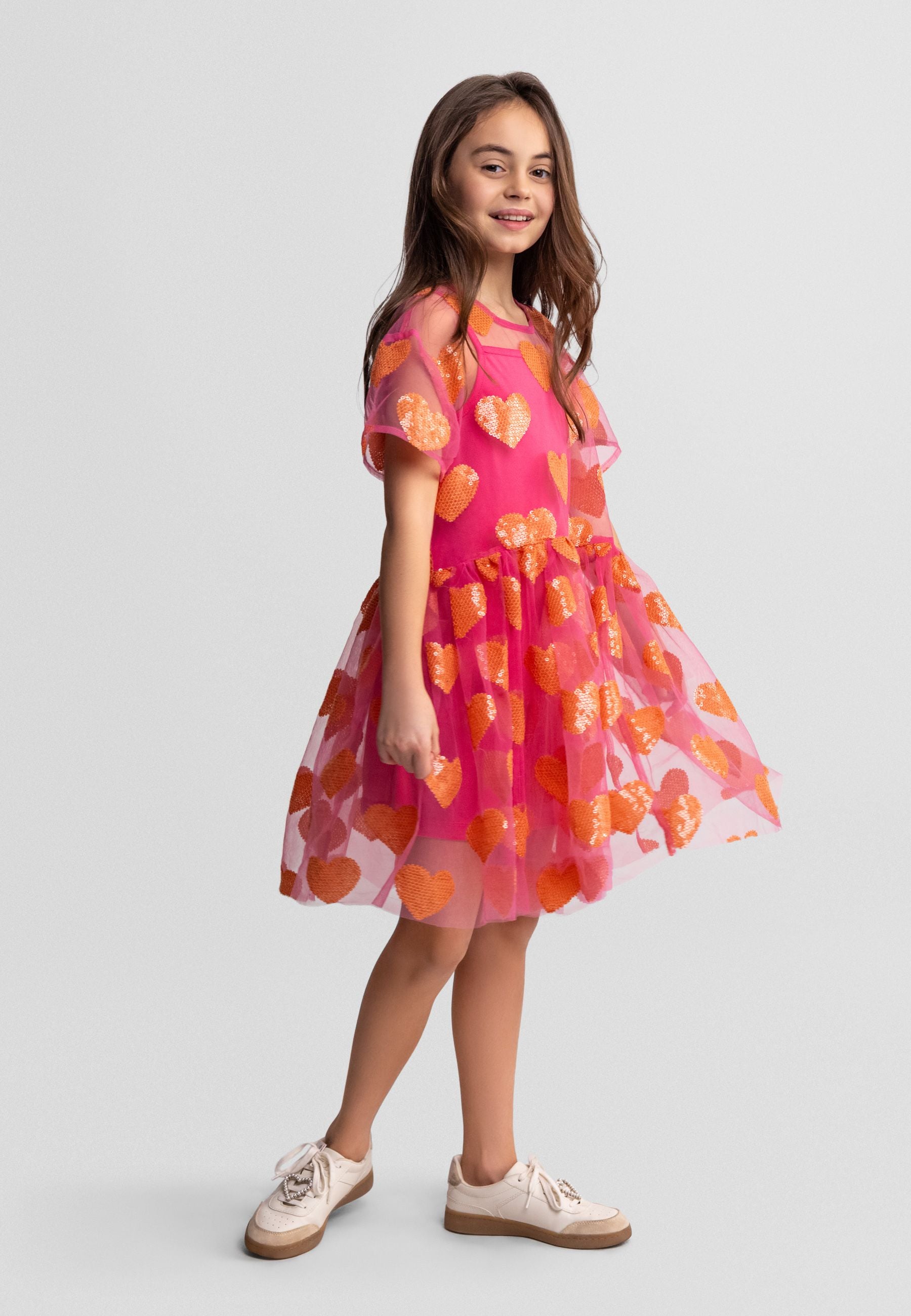 Young girl wearing pink tulle dress with sequin hearts, smiling at camera, standing against light background wearing beige sneakers
