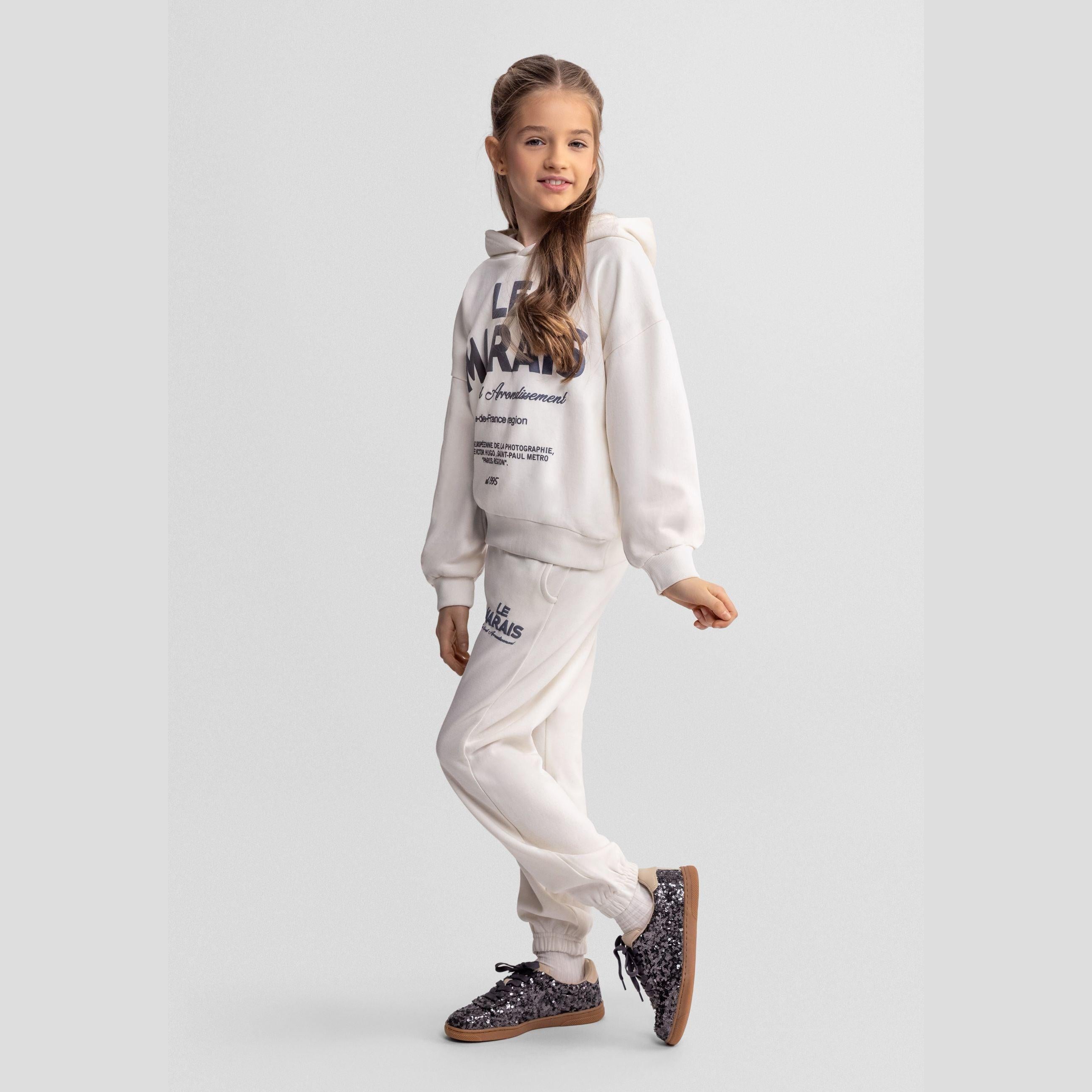Girl wearing off white tracksuit set with hooded sweatshirt and joggers, Le Marais text print visible, standing pose against light grey background