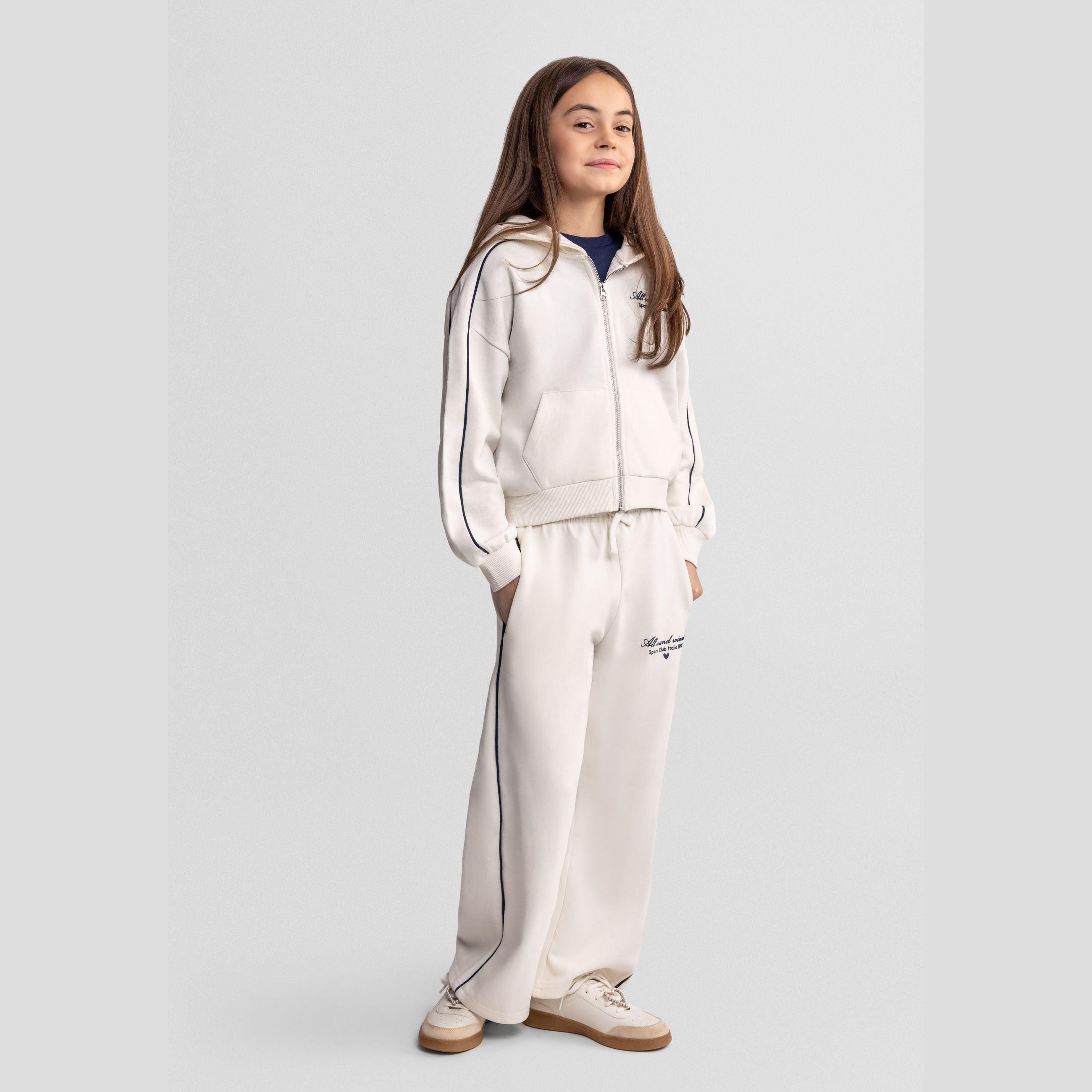 Girl wearing beige tracksuit set with zip hoodie and wide-leg trousers, navy contrast piping, standing against light grey background