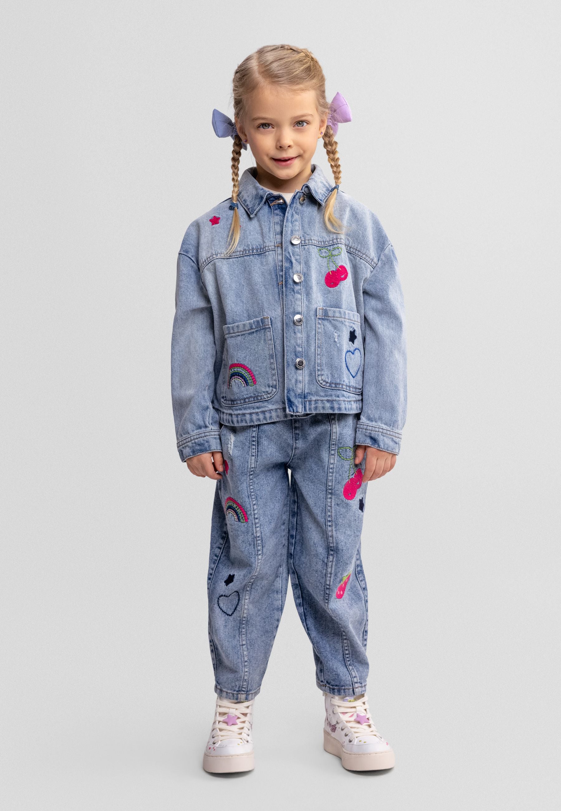Young girl wearing blue denim barrel leg jeans with embroidered details and matching jacket, standing against white background with braided hair and hair clips