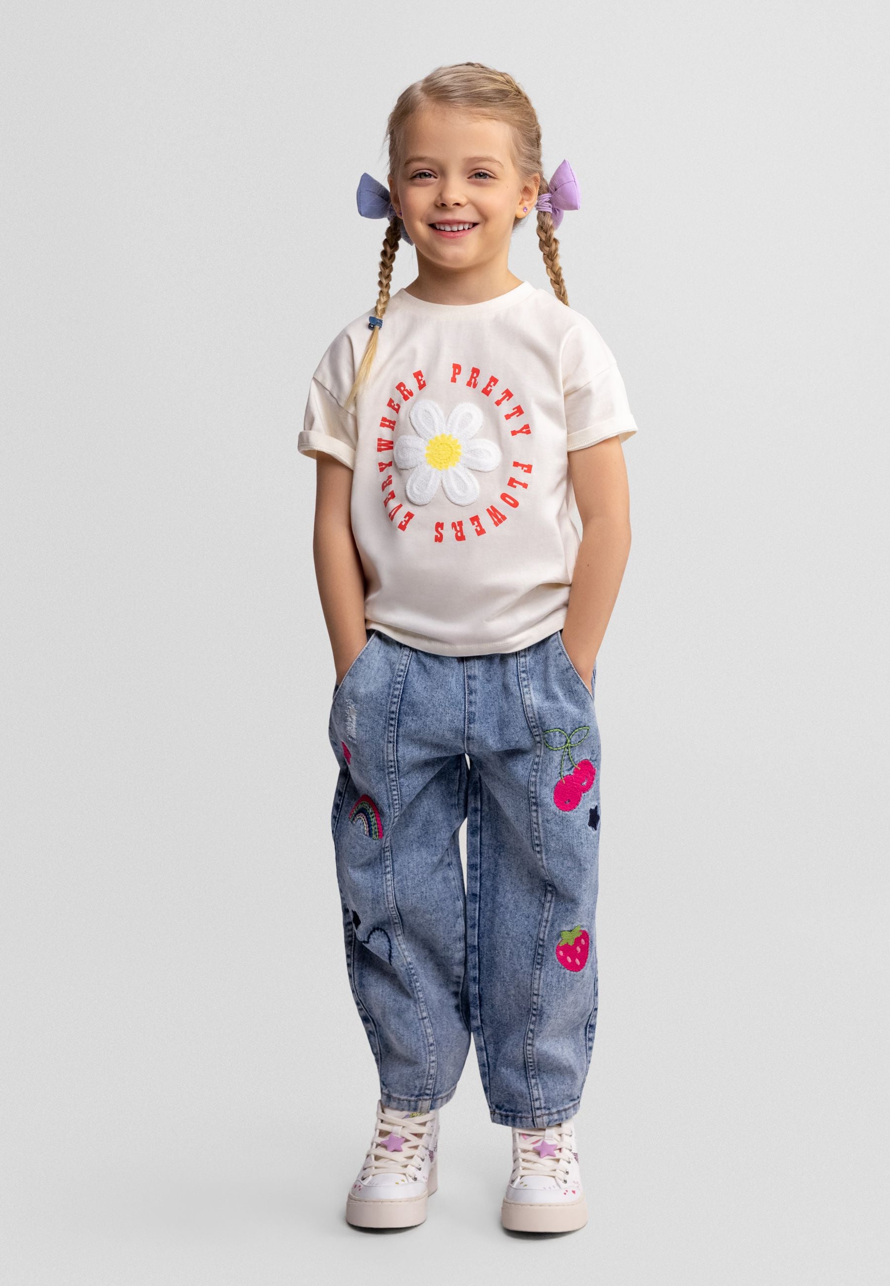 Close-up flat lay of blue denim barrel leg jeans showing elastic waistband with pink drawstring and embroidered details including rainbows and fruits