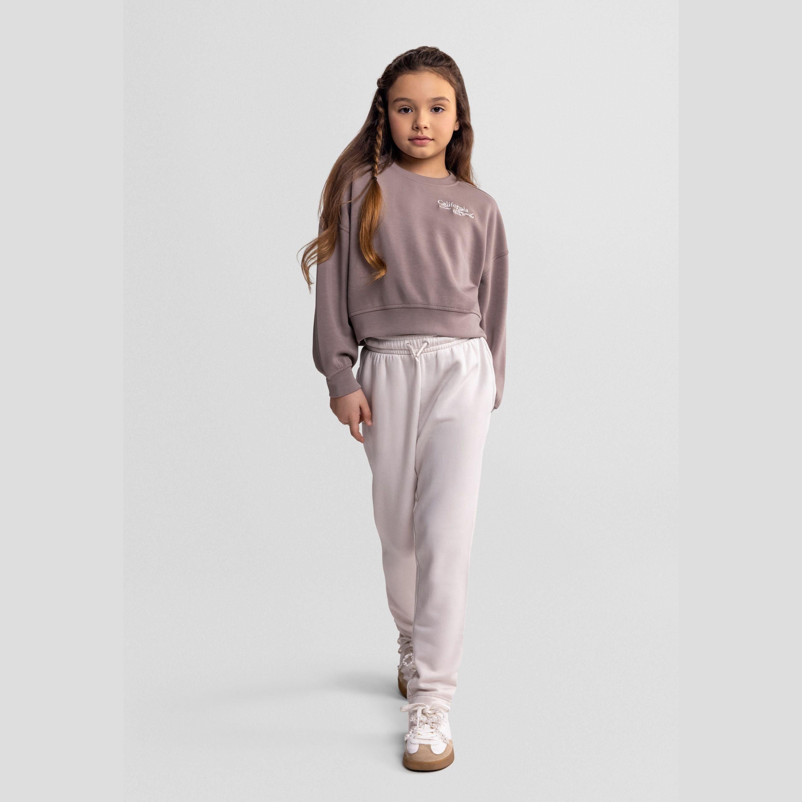 Young girl wearing brown crew neck sweatshirt with white joggers against light grey background, full body studio shot