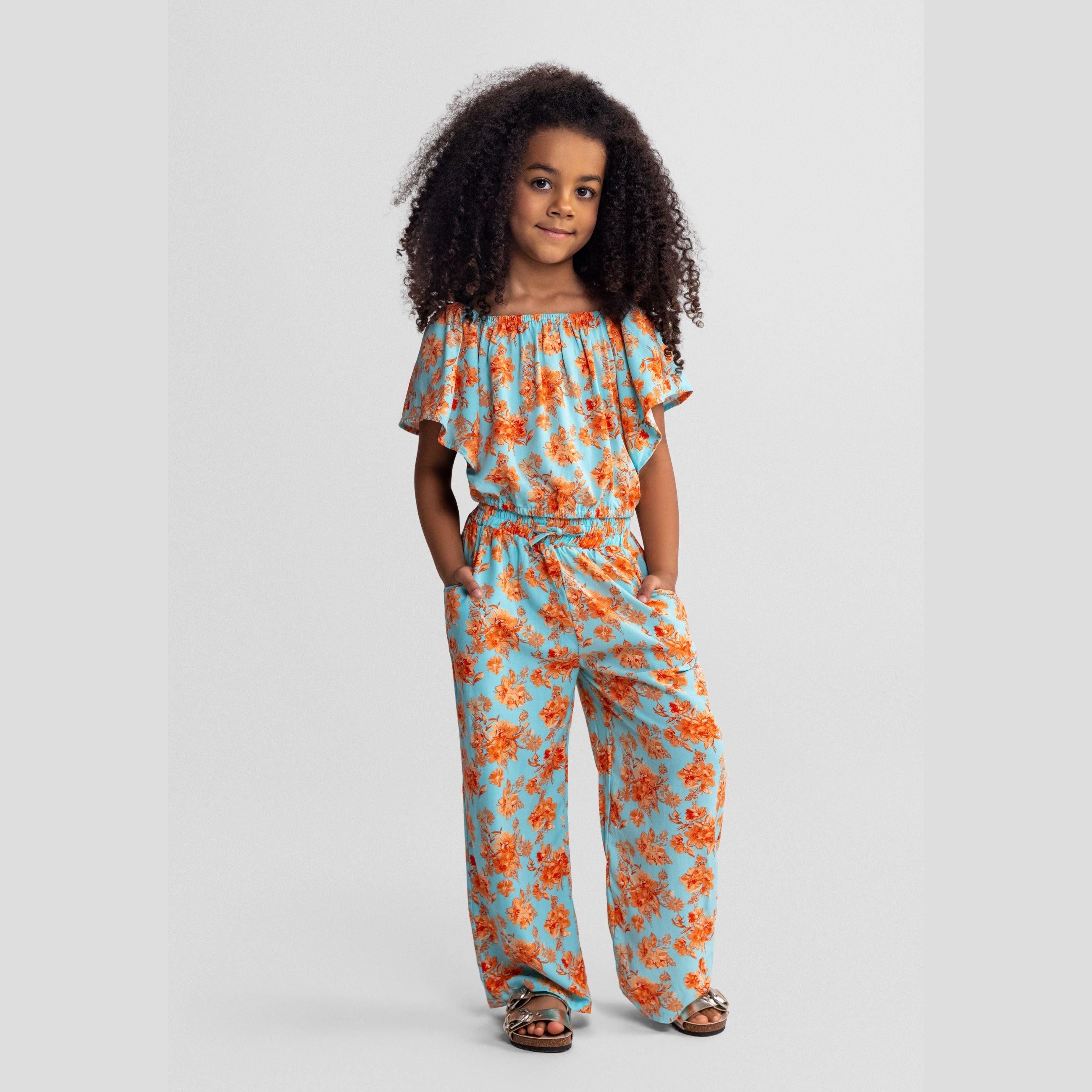 Young girl wearing light blue viscose jumpsuit with orange floral print, featuring off-shoulder design and wide-leg trousers, standing against white background