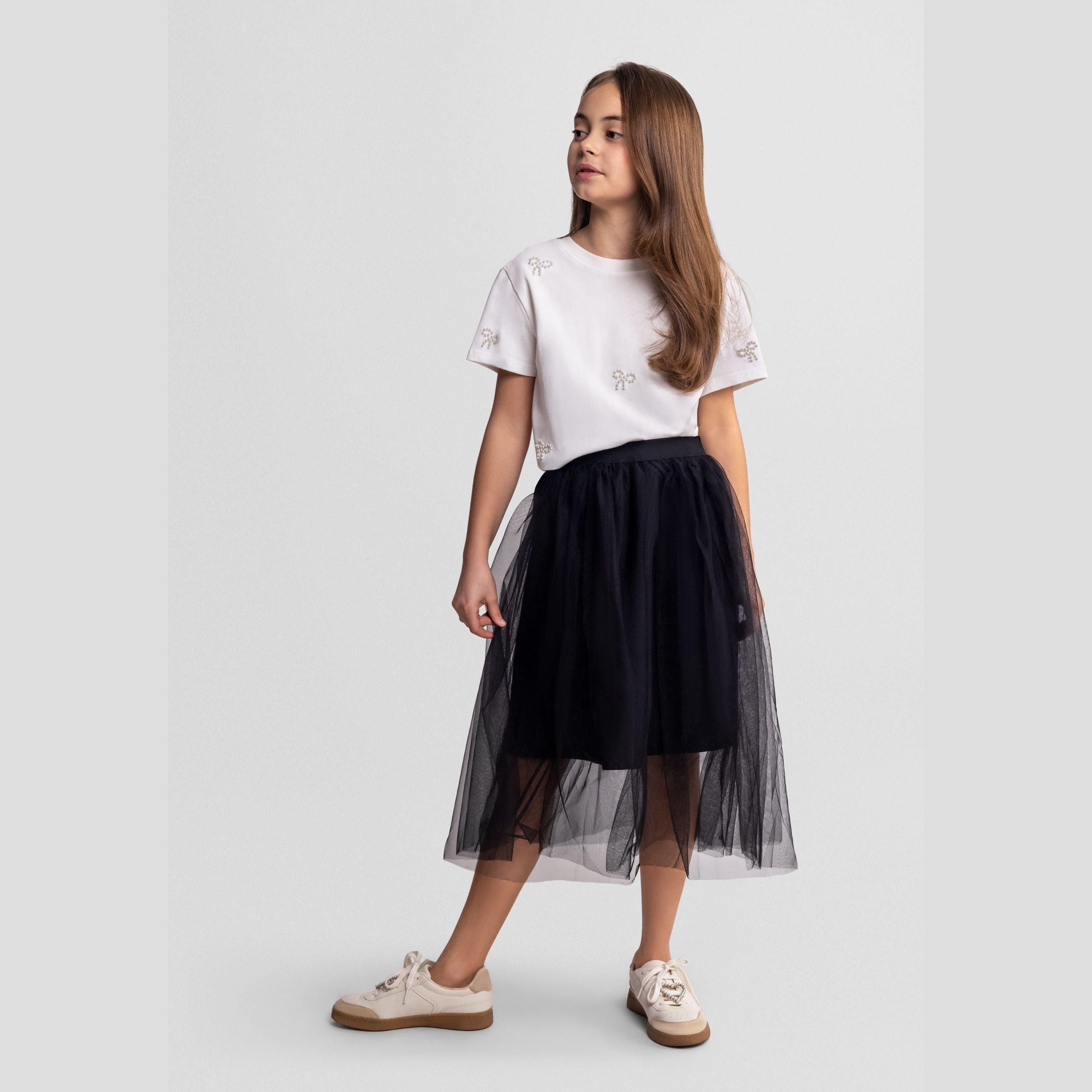 Young girl wearing black pleated tulle maxi skirt with white embellished t-shirt, standing against light grey background