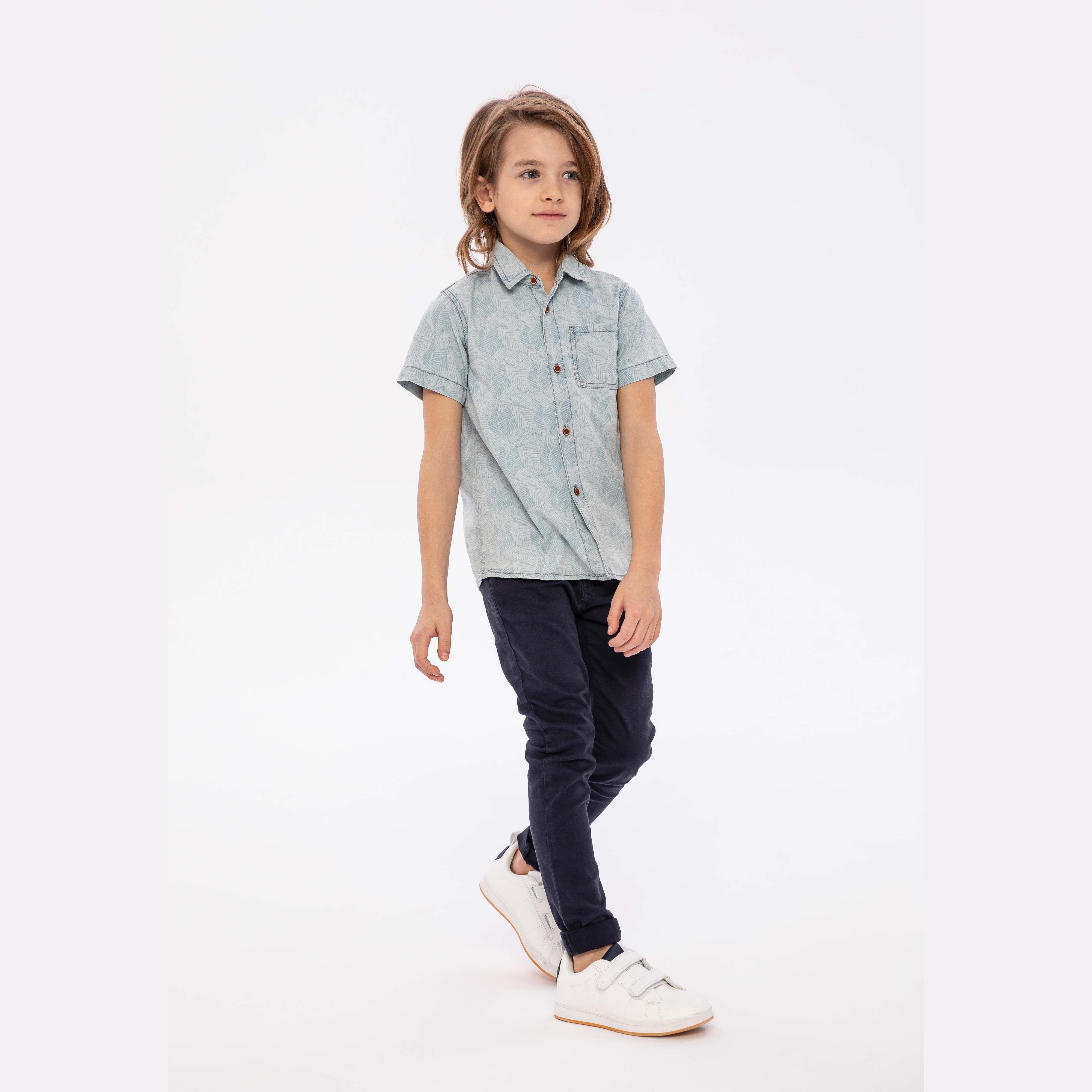 Young boy wearing light blue bleached denim shirt with dark jeans against white studio background, full body casual pose