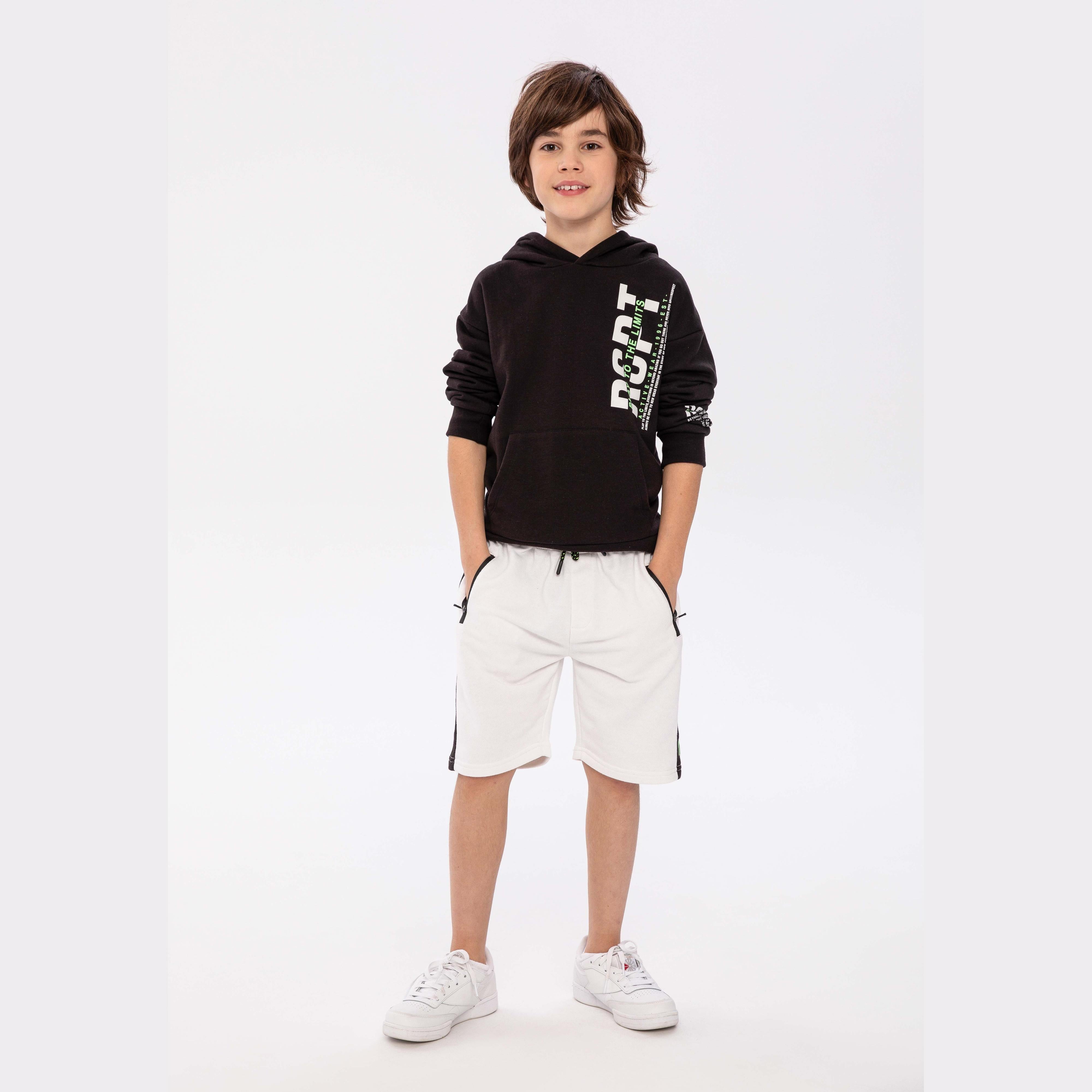 Boy wearing black fleece hoodie with white branded print and light grey shorts, standing against white studio background