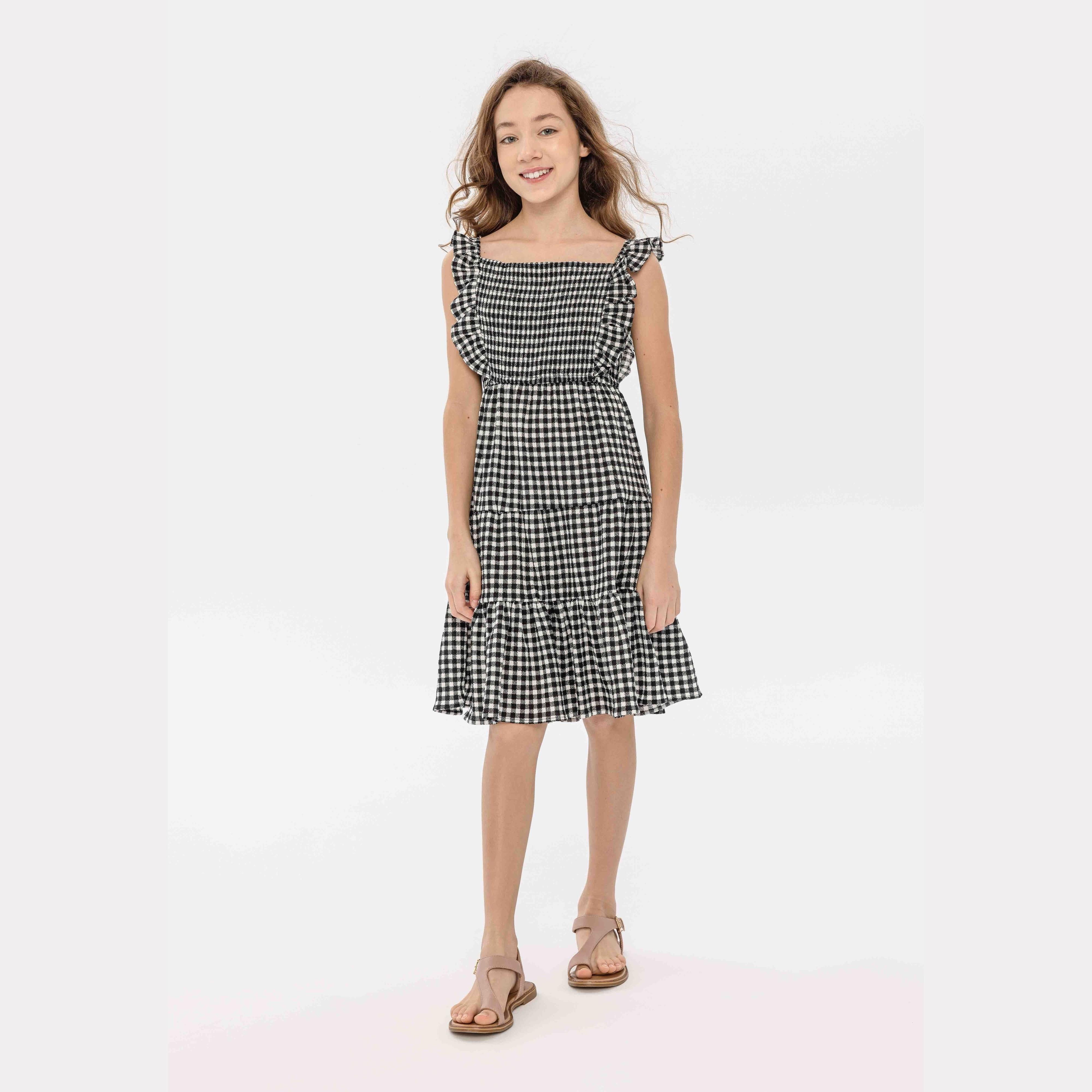 Young girl wearing black and white gingham tiered dress with flutter sleeves, standing against light background wearing brown sandals