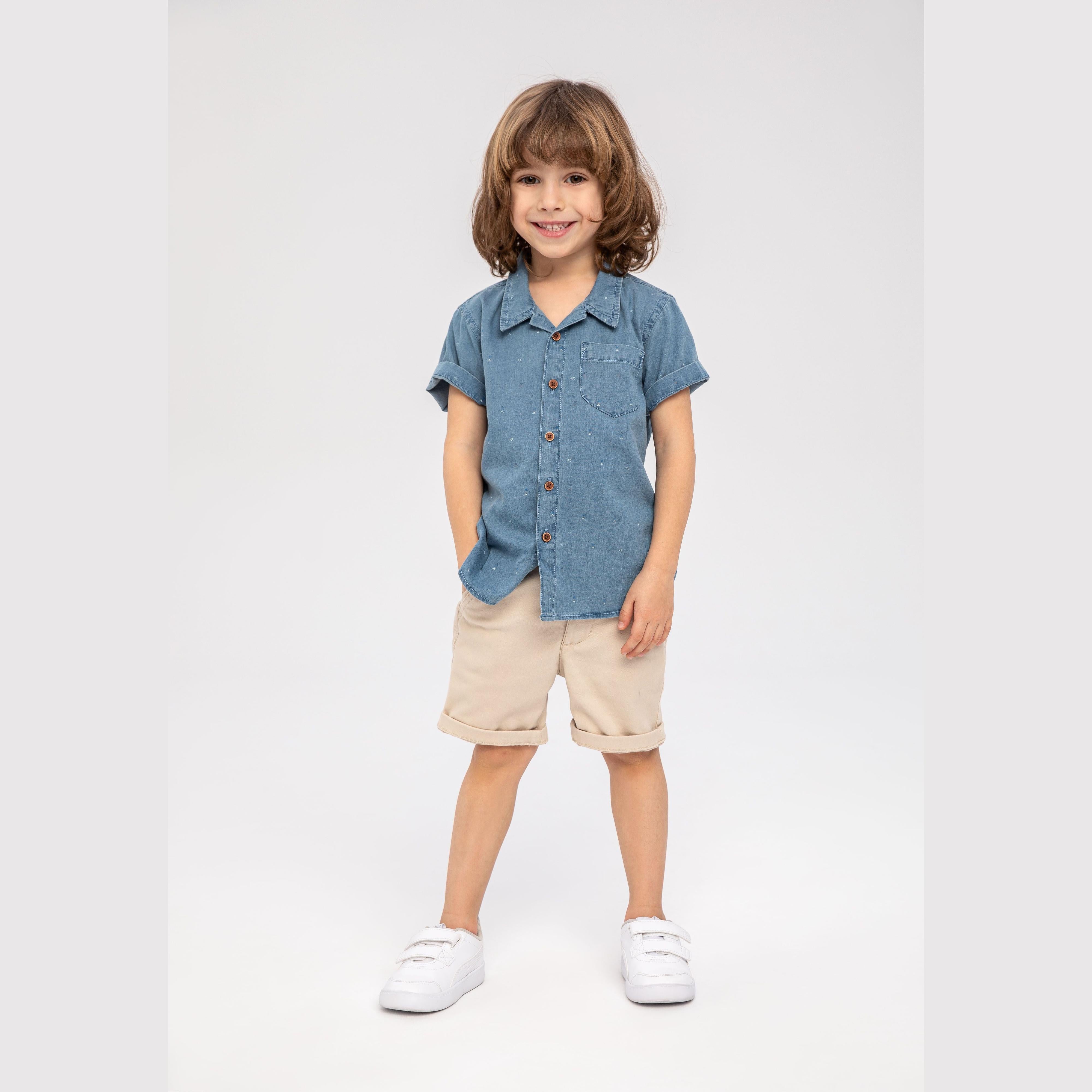 Young child with brown bob haircut wearing light blue printed denim shirt with beige shorts and white sneakers on light background