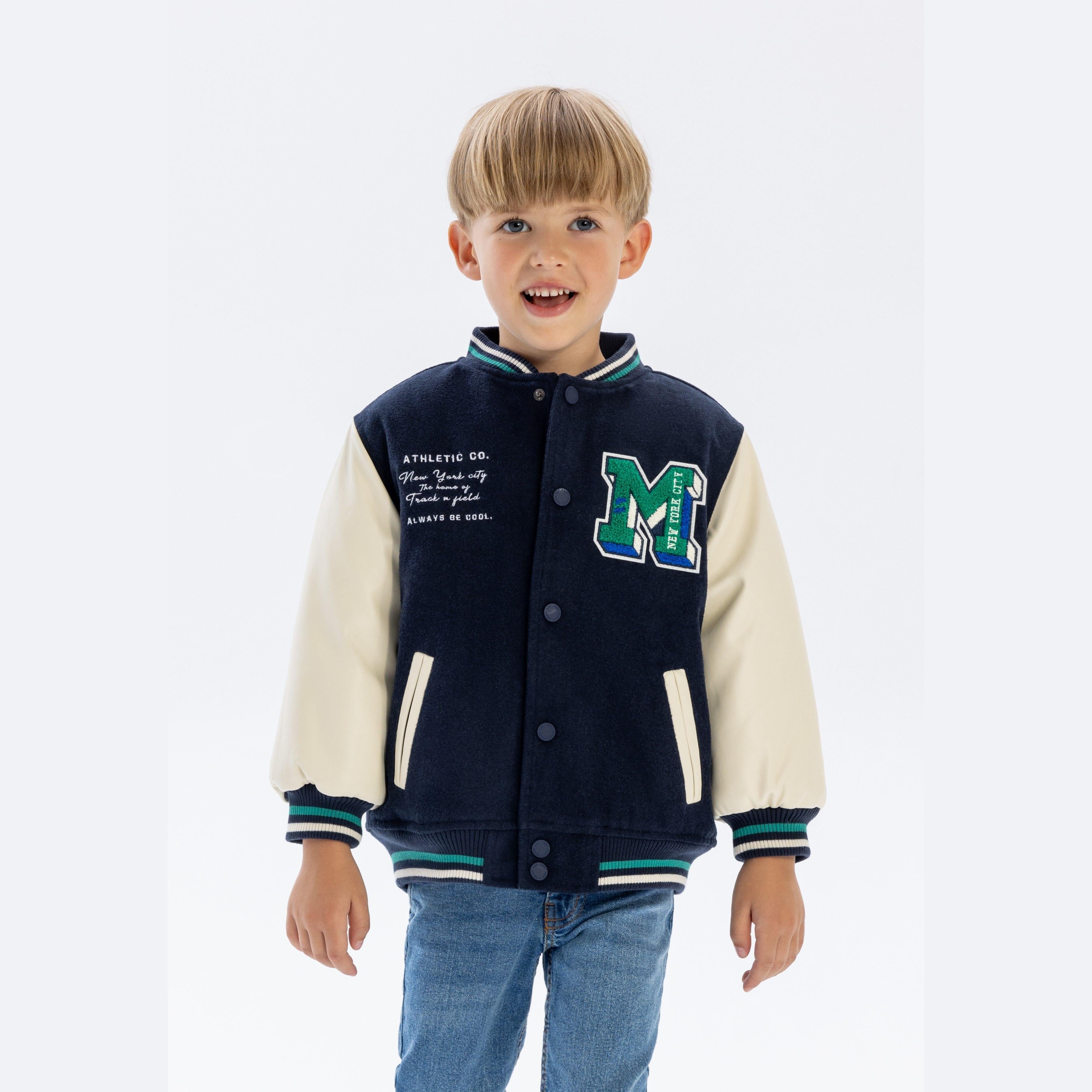 Young boy wearing navy and cream baseball jacket with green M appliqué, smiling against white studio background