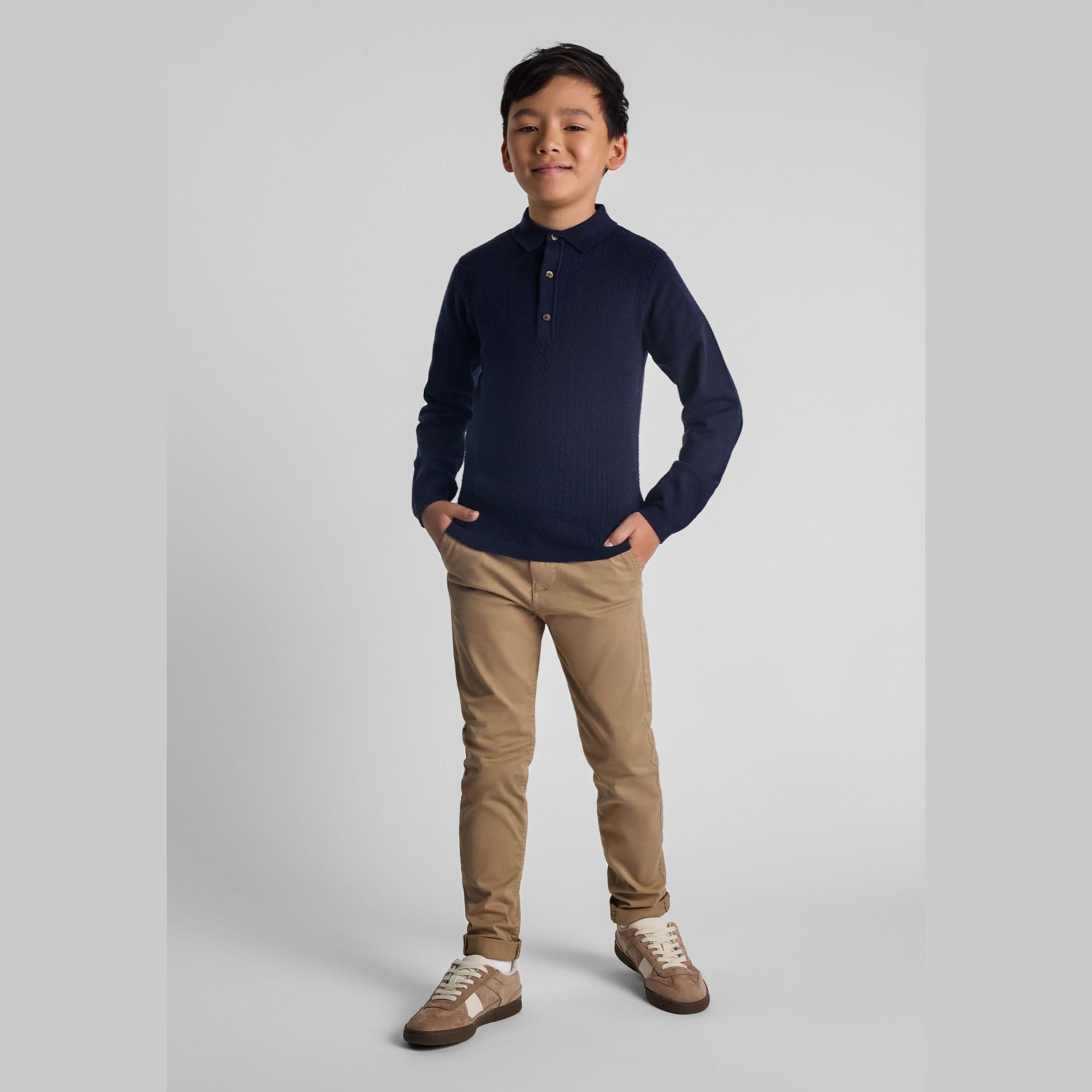 Young boy wearing navy blue long-sleeve polo shirt and beige chino trousers, standing confidently against white studio background